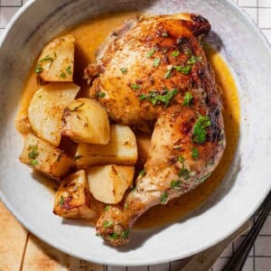 A plate with a roasted chicken leg quarter, golden potatoes, and sauce, garnished with herbs. The plate sits on a white checked tablecloth, with bread, a fork, and a knife partially visible beside it.
