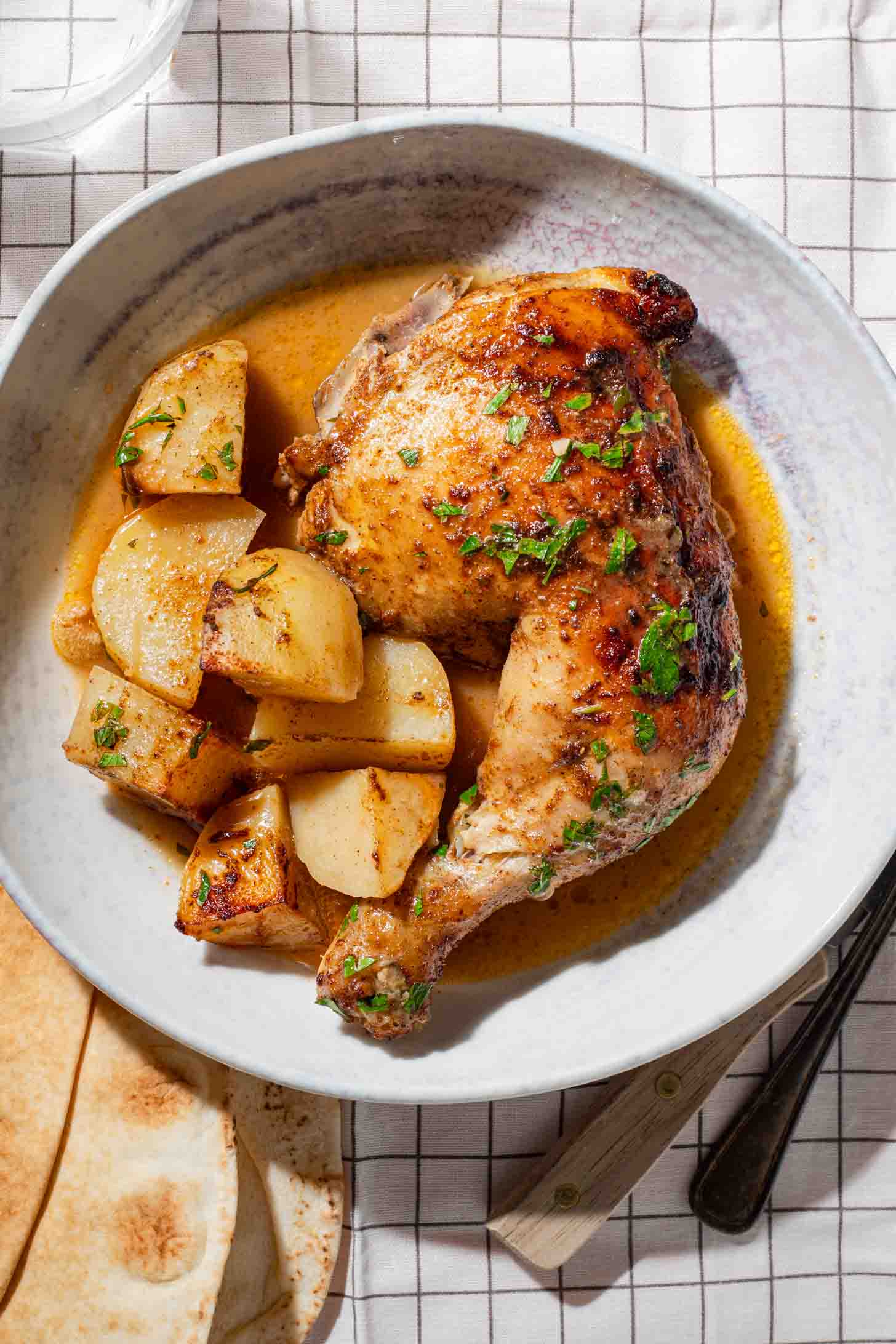 A plate with a roasted chicken leg quarter, golden potatoes, and sauce, garnished with herbs. The plate sits on a white checked tablecloth, with bread, a fork, and a knife partially visible beside it.