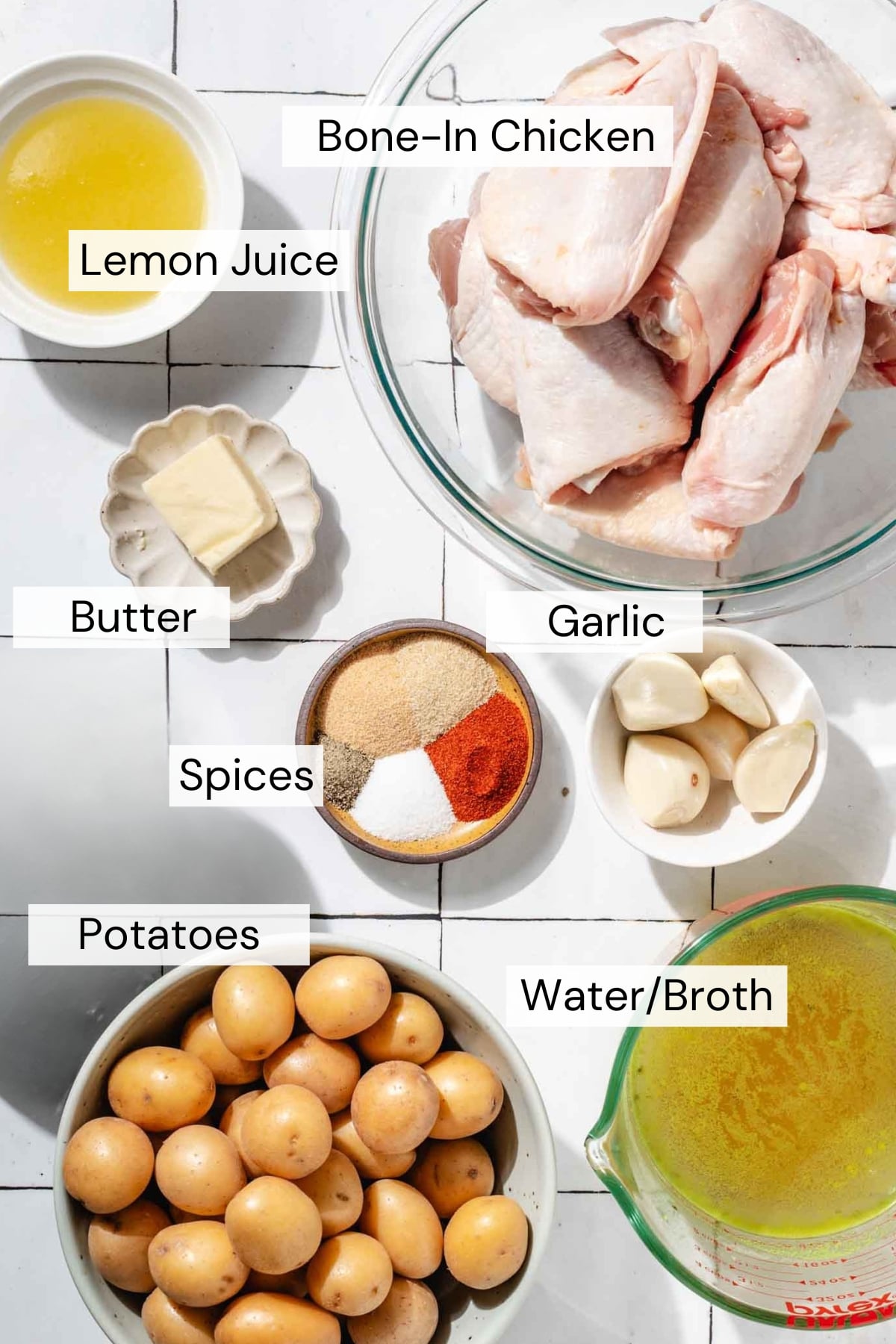 A flat lay of labeled ingredients including bone-in chicken, lemon juice, butter, spices, garlic, potatoes, and a measuring cup of water or broth on a white surface.