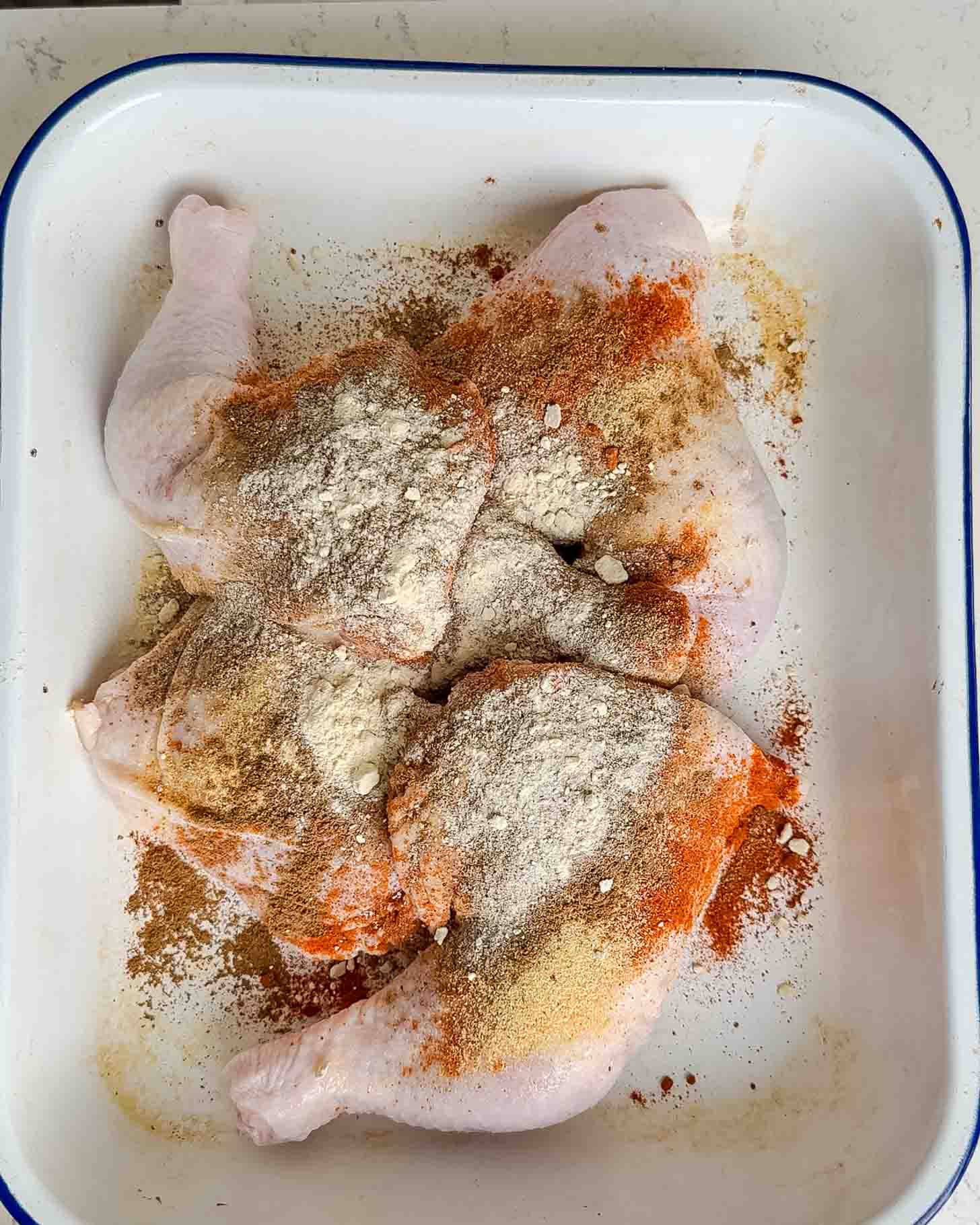 Raw chicken legs in a white tray, sprinkled with various spices and flour, ready for seasoning or marinating.