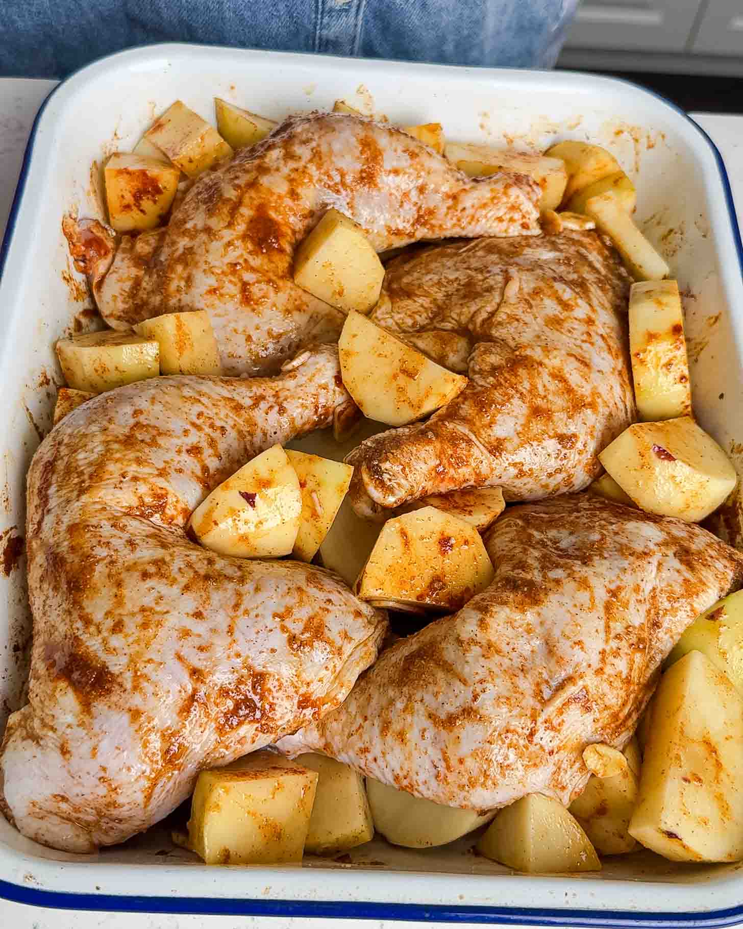 Raw, seasoned chicken legs with spices arranged in a baking dish, surrounded by chopped potato pieces, ready to be cooked.