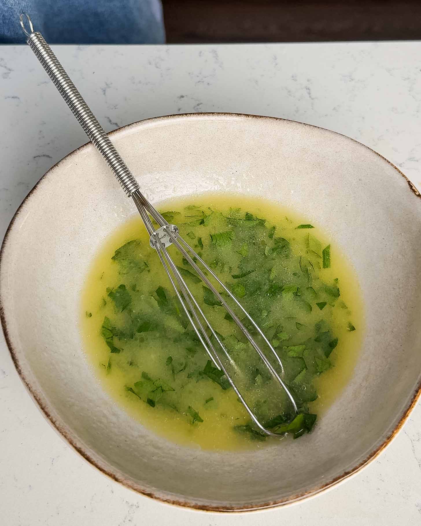 A white bowl with a metal whisk resting inside, containing a green herb vinaigrette or salad dressing on a white countertop.
