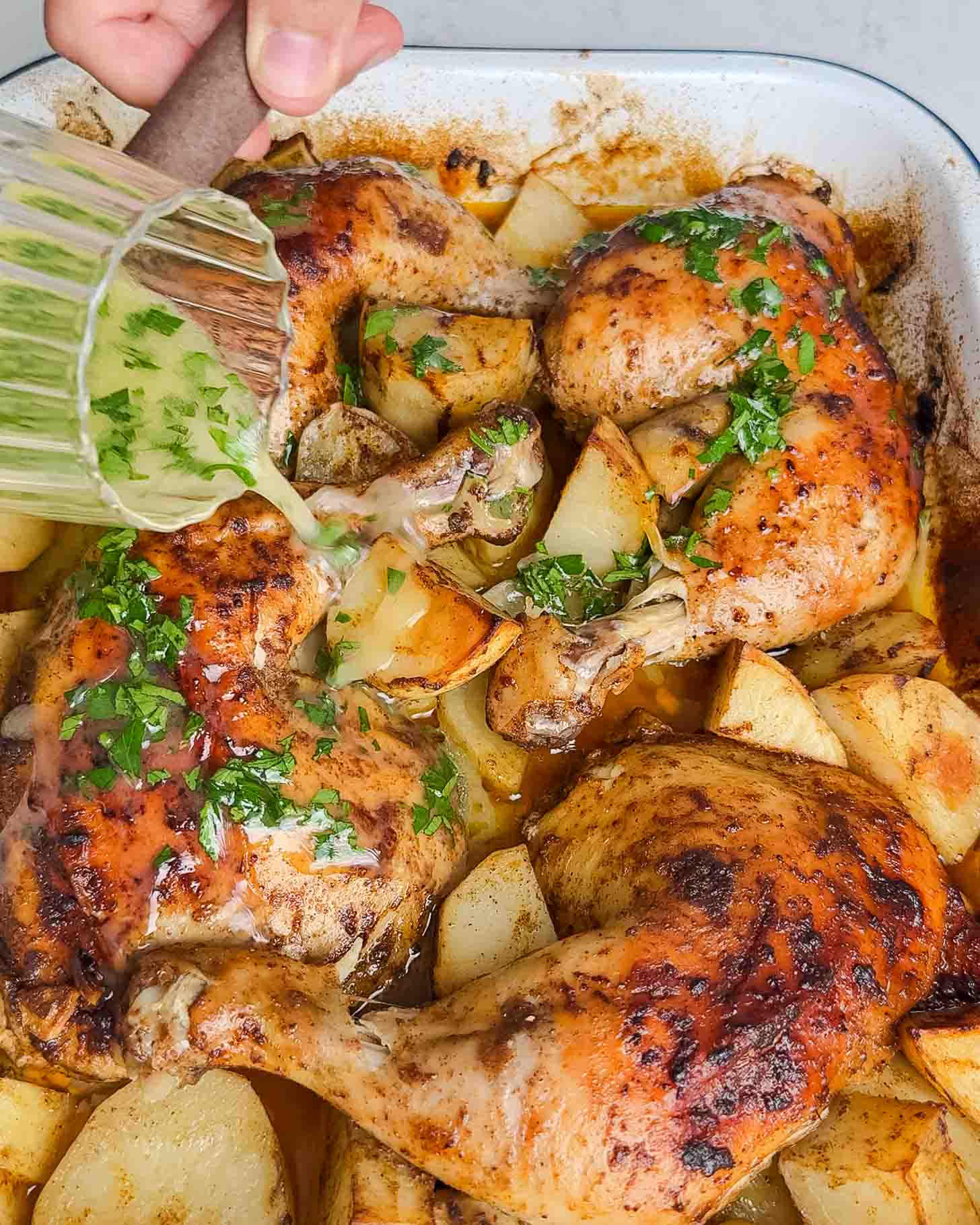 A hand pours a green herb sauce over roasted chicken legs and potato wedges in a baking dish. The dish is garnished with chopped fresh herbs.