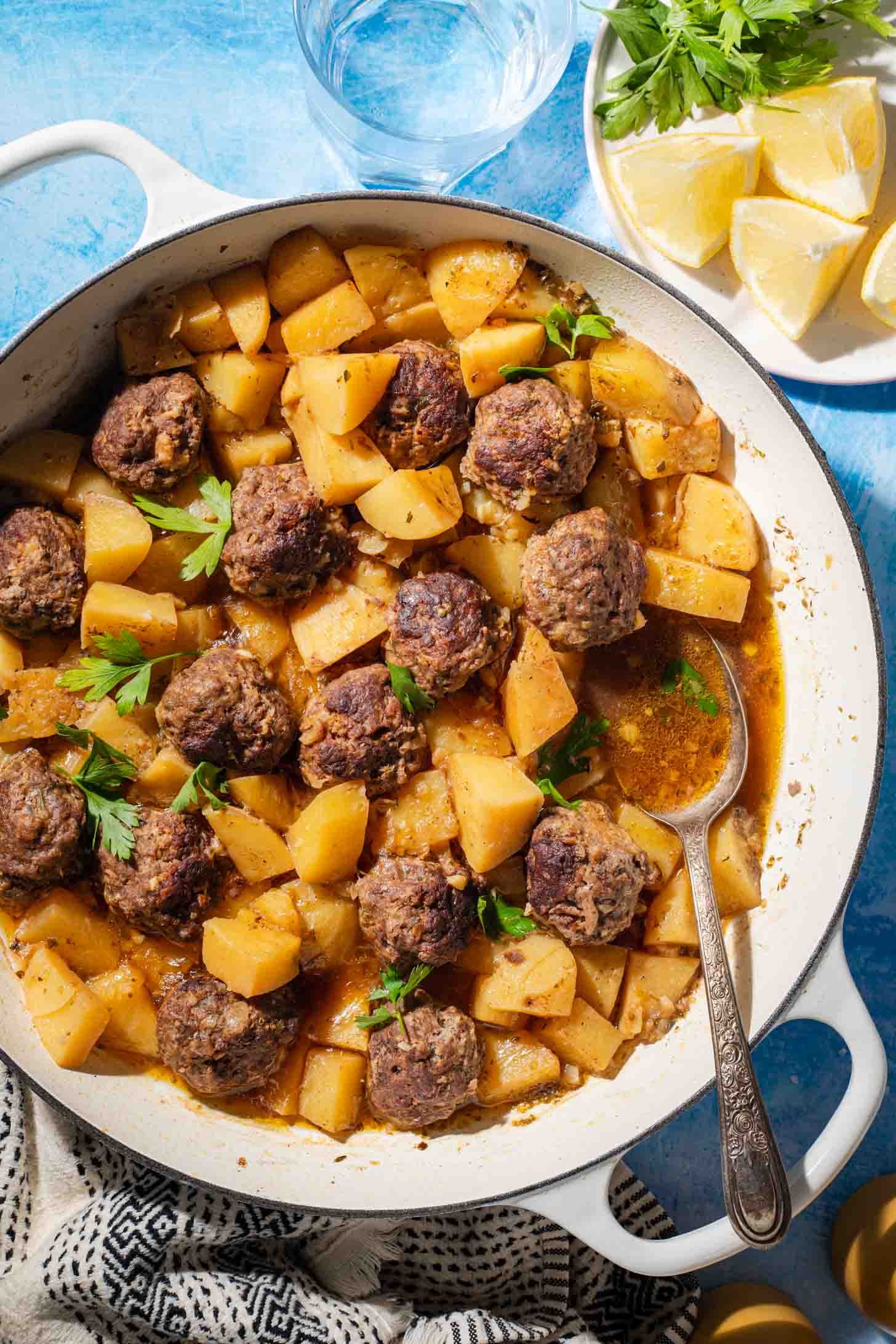 A white pan filled with meatballs, potatoes, and sauce, garnished with parsley. A spoon rests in the pan. Nearby are lemon wedges, parsley, a glass of water, and a black-and-white towel. Blue surface underneath.