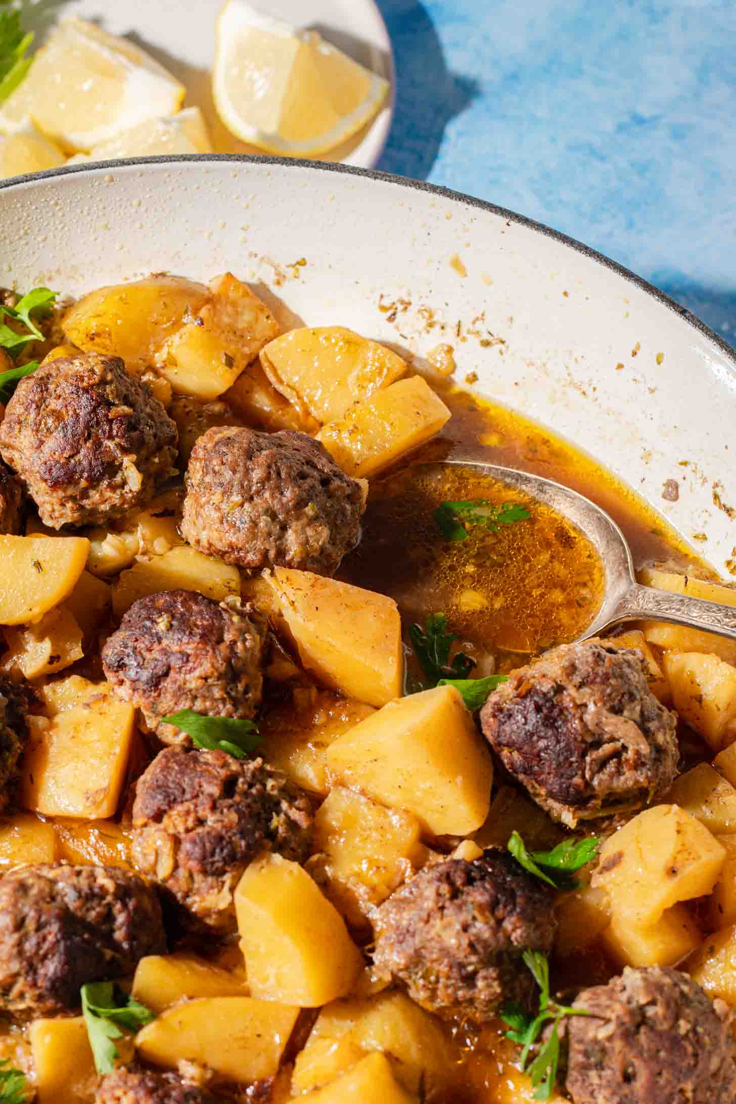 A skillet filled with browned meatballs, cubed potatoes, and fresh herbs in a savory broth, with lemon wedges on a plate in the background.