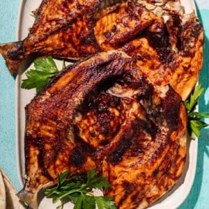 Grilled fish pieces with golden, crispy skin on a white platter, garnished with fresh parsley, on a light blue background.