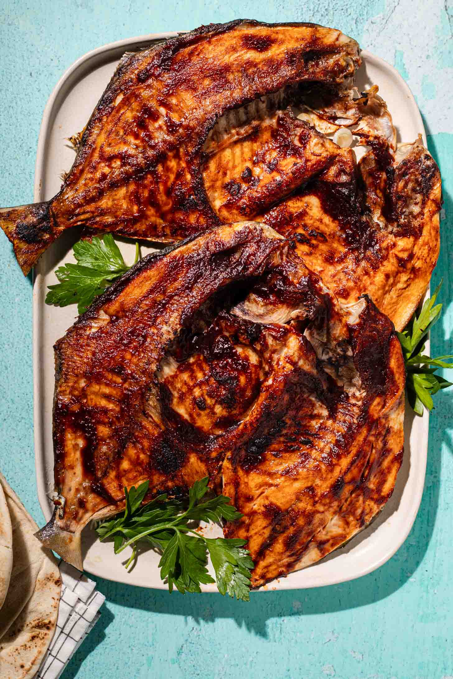 Grilled fish pieces with golden, crispy skin on a white platter, garnished with fresh parsley, on a light blue background.