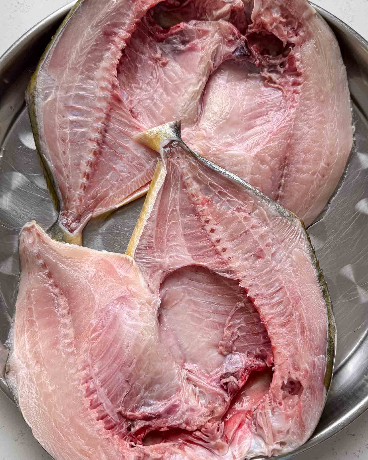 Two raw, split fish laid open on a round metal tray, showing pink flesh and bones. The fish are arranged to display their inner sides and occupy most of the tray.