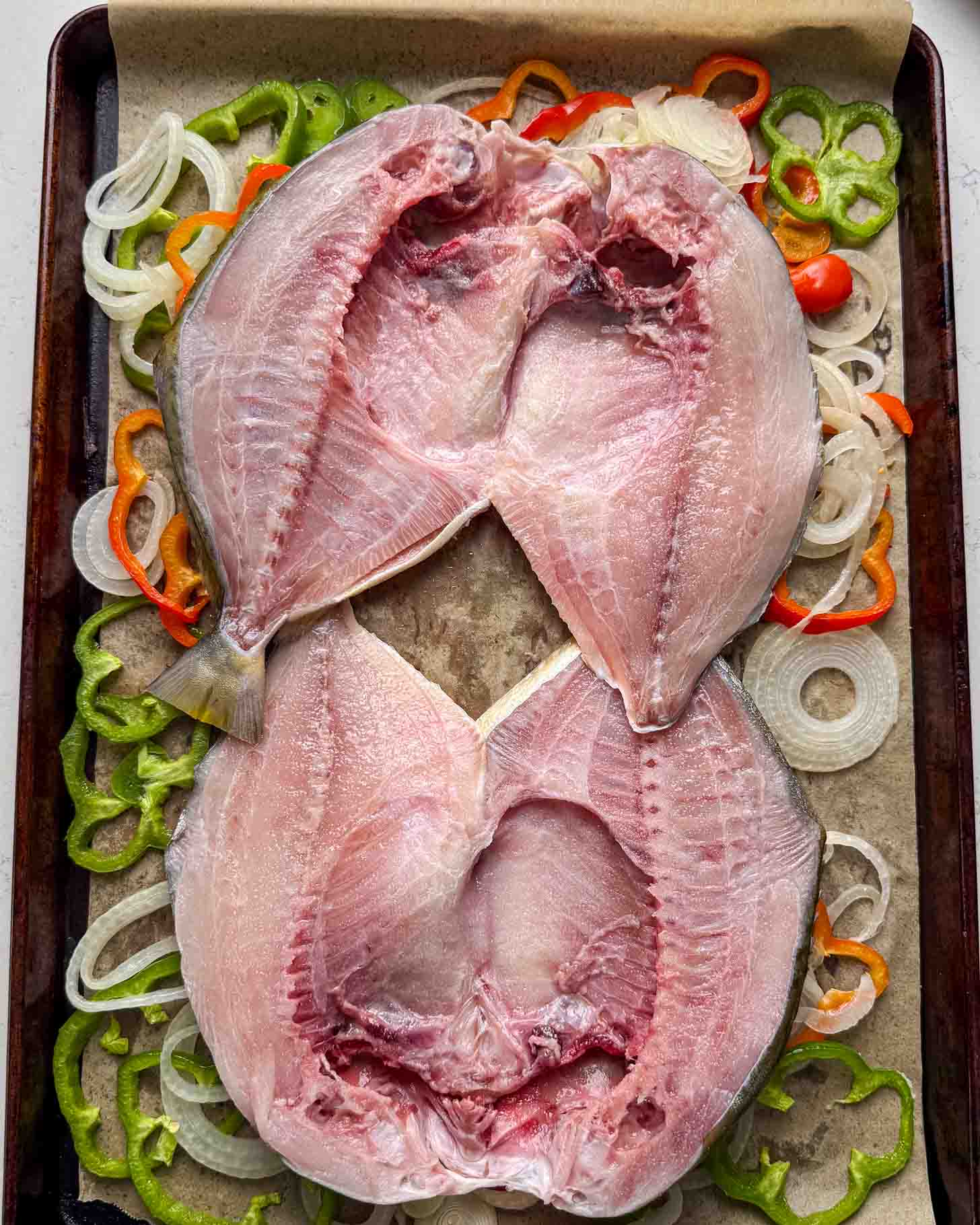 Two split, raw fish laid open on a baking tray lined with parchment paper, surrounded by sliced onions and green and orange bell peppers.