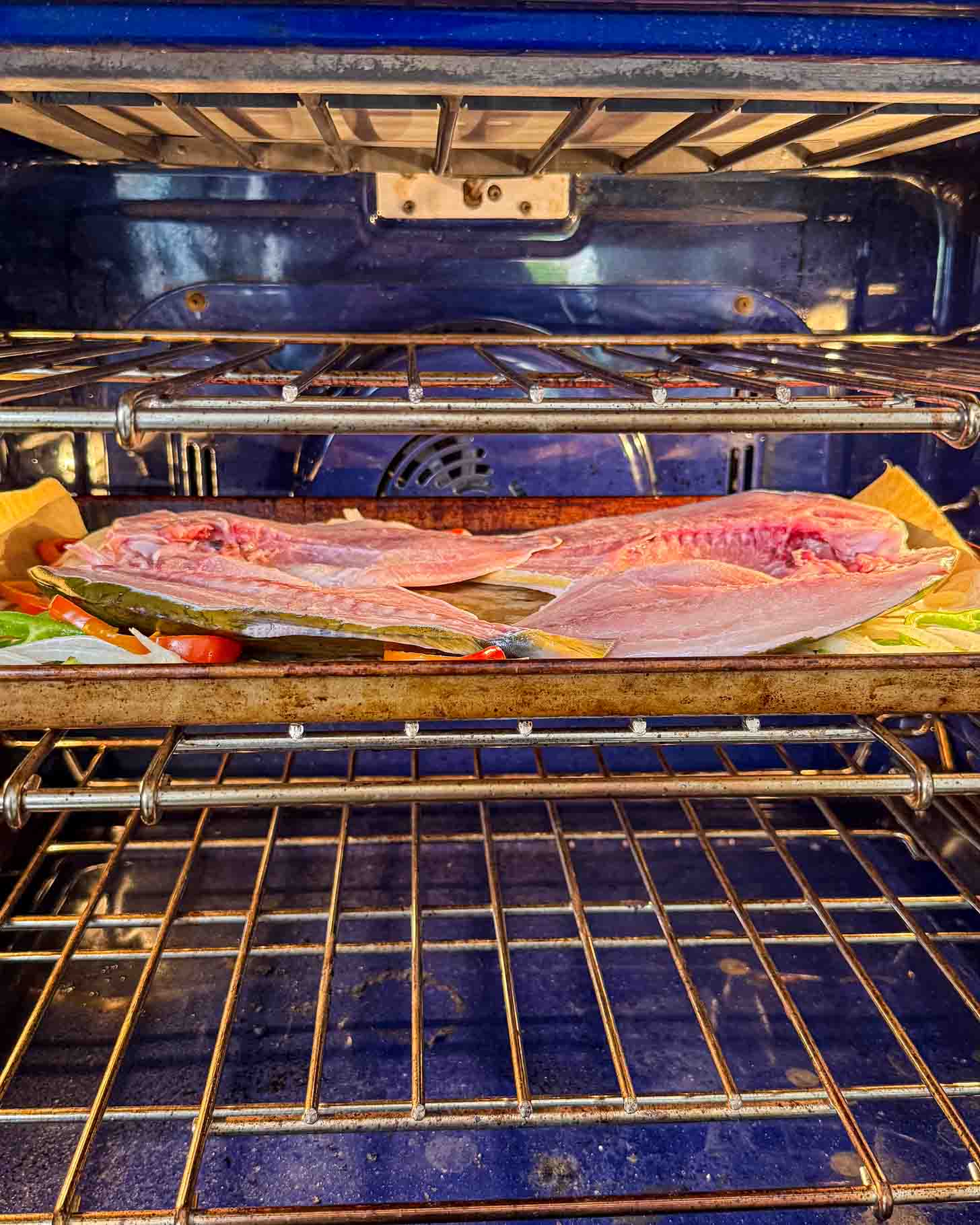 A whole fish, split open and laid flat on a baking tray, is placed on the middle rack of a blue oven, ready to be cooked.