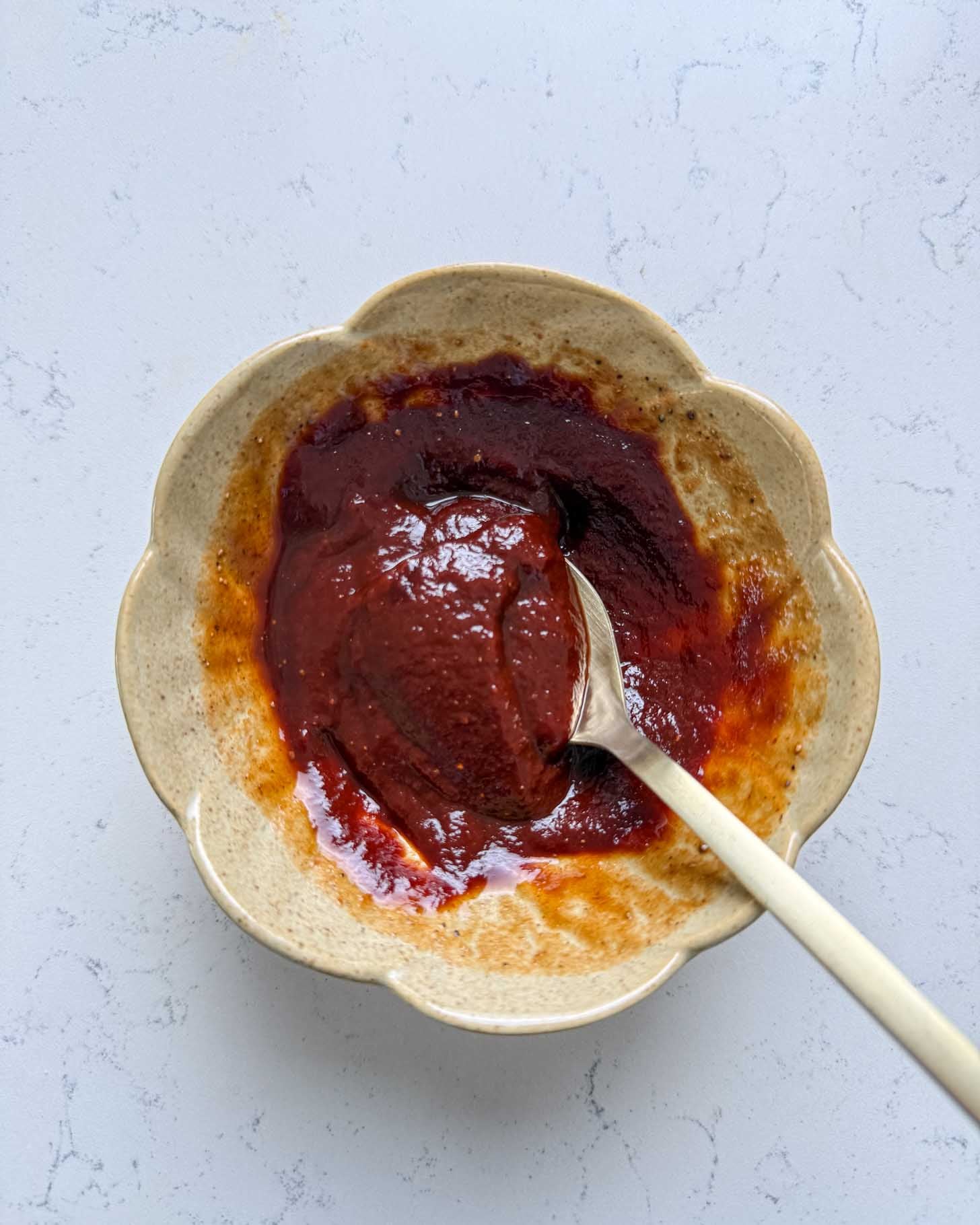 A beige, scalloped bowl with thick, dark reddish-brown sauce and a spoon rests inside, on a light, textured surface.