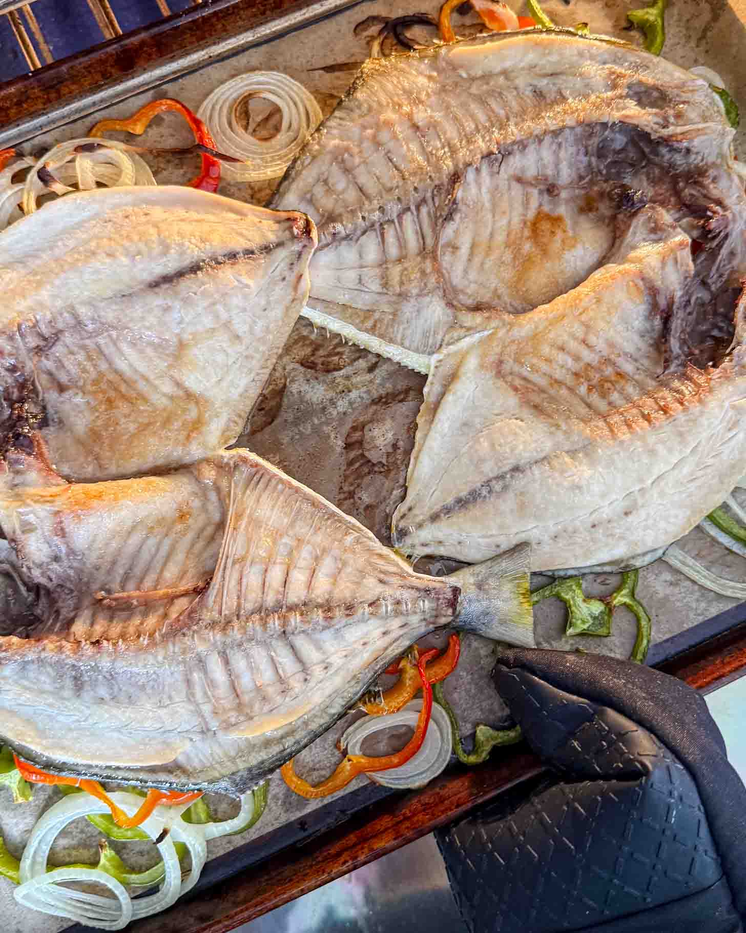 Three cleaned fish laid open on a baking tray atop sliced onions and bell peppers, with a gloved hand holding the tray.