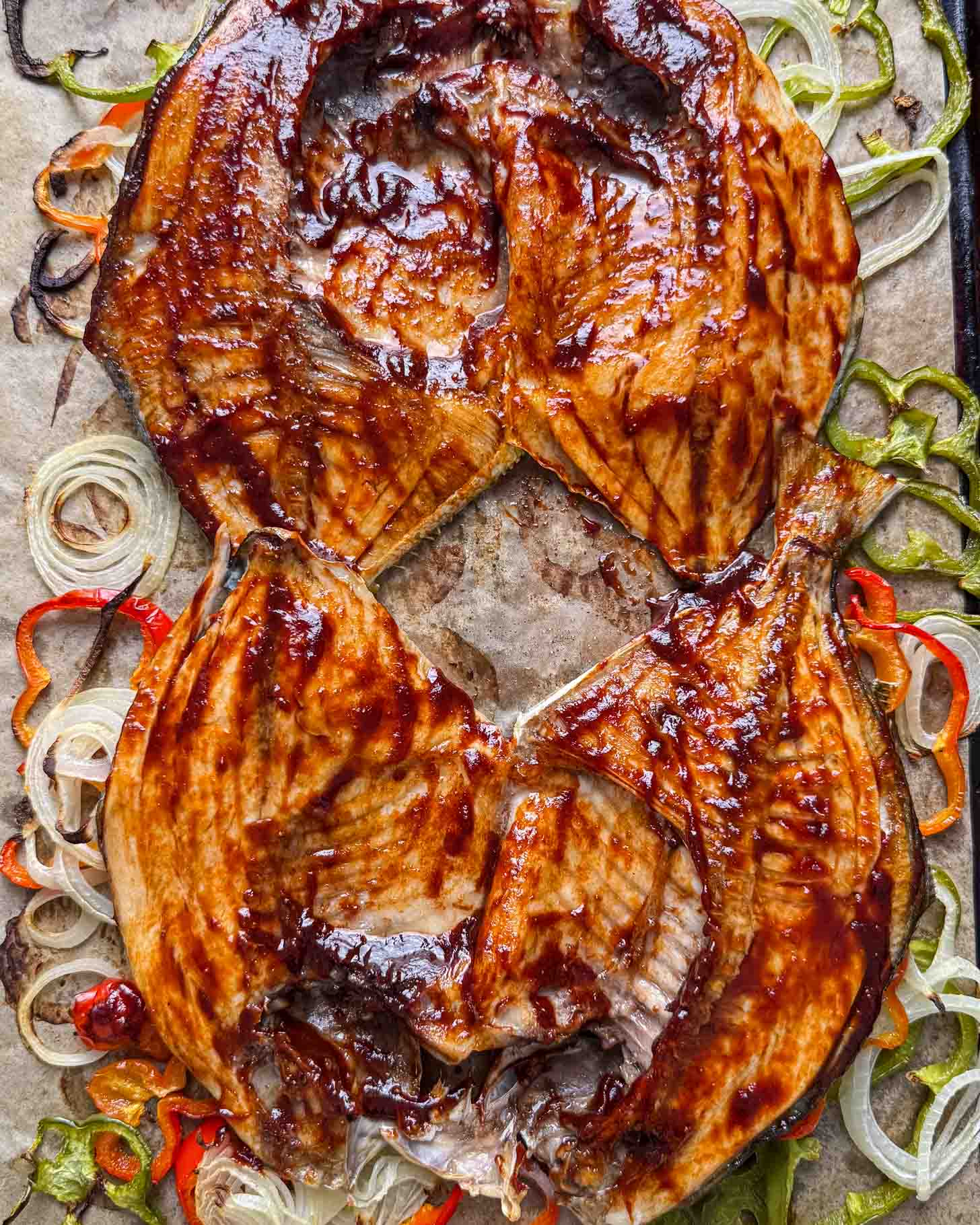 Grilled, butterflied fish coated in barbecue sauce on a bed of sliced onions and bell peppers, all arranged on parchment paper.