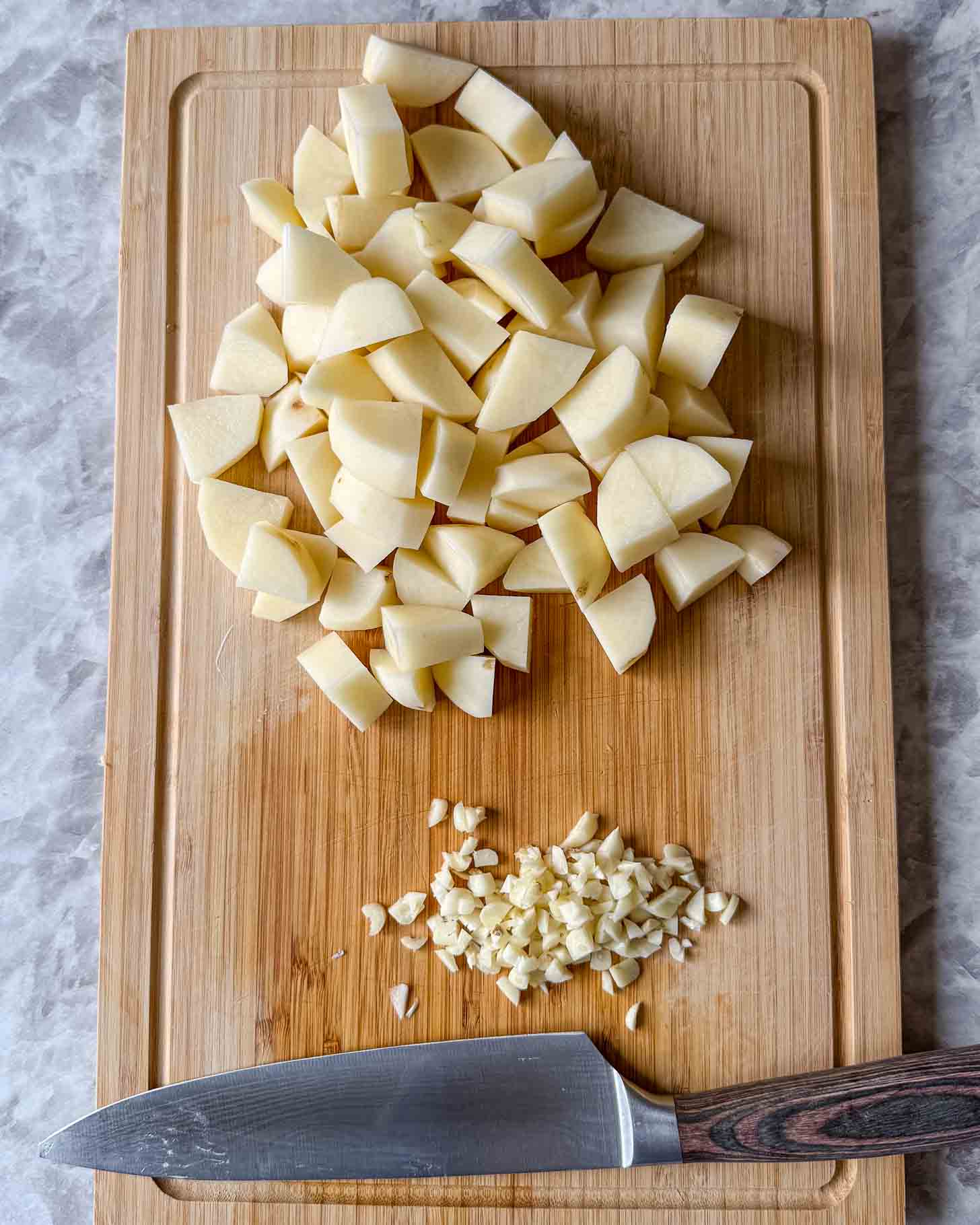 Diced potatoes and minced garlic are on a wooden cutting board, with a large kitchen knife placed below them on a marble surface.