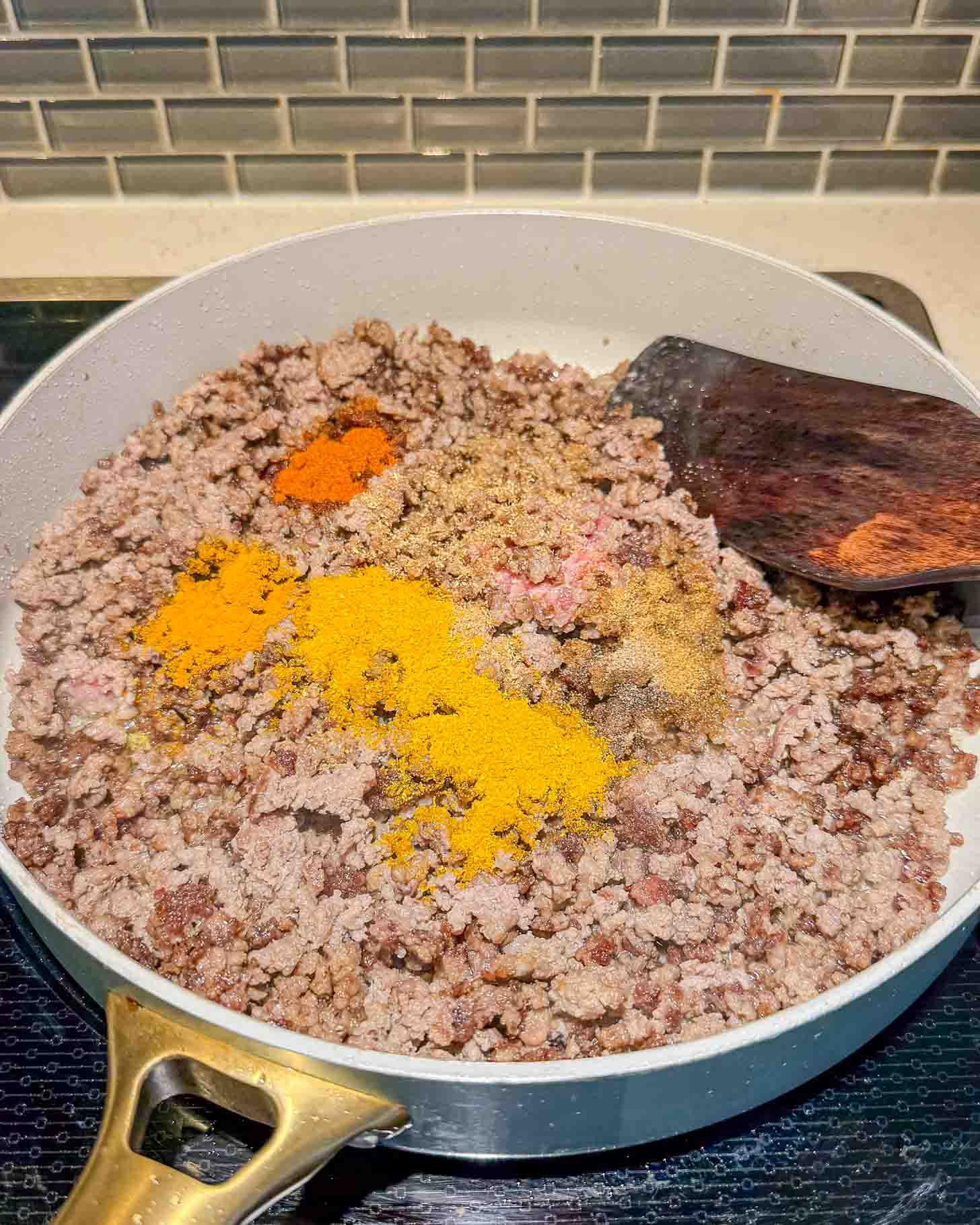 Ground beef cooking in a white skillet on a stove, topped with various colorful spices. A wooden spatula rests on the side of the pan, and a tiled backsplash is visible in the background.