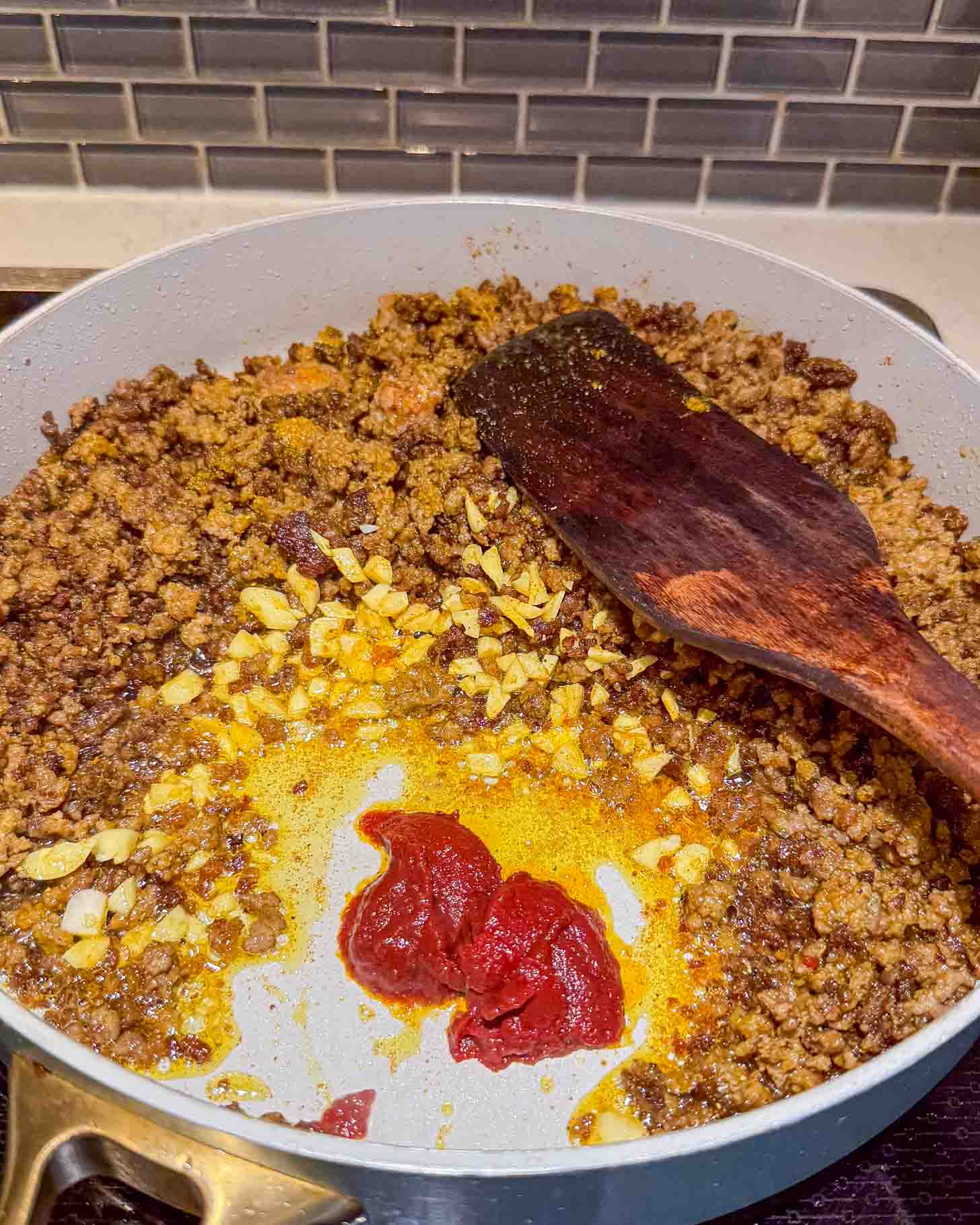 Ground meat is being sautéed with chopped onions, garlic, and tomato paste in a large white pan on a stove. A wooden spatula rests to the side, and a gray tile backsplash is visible in the background.