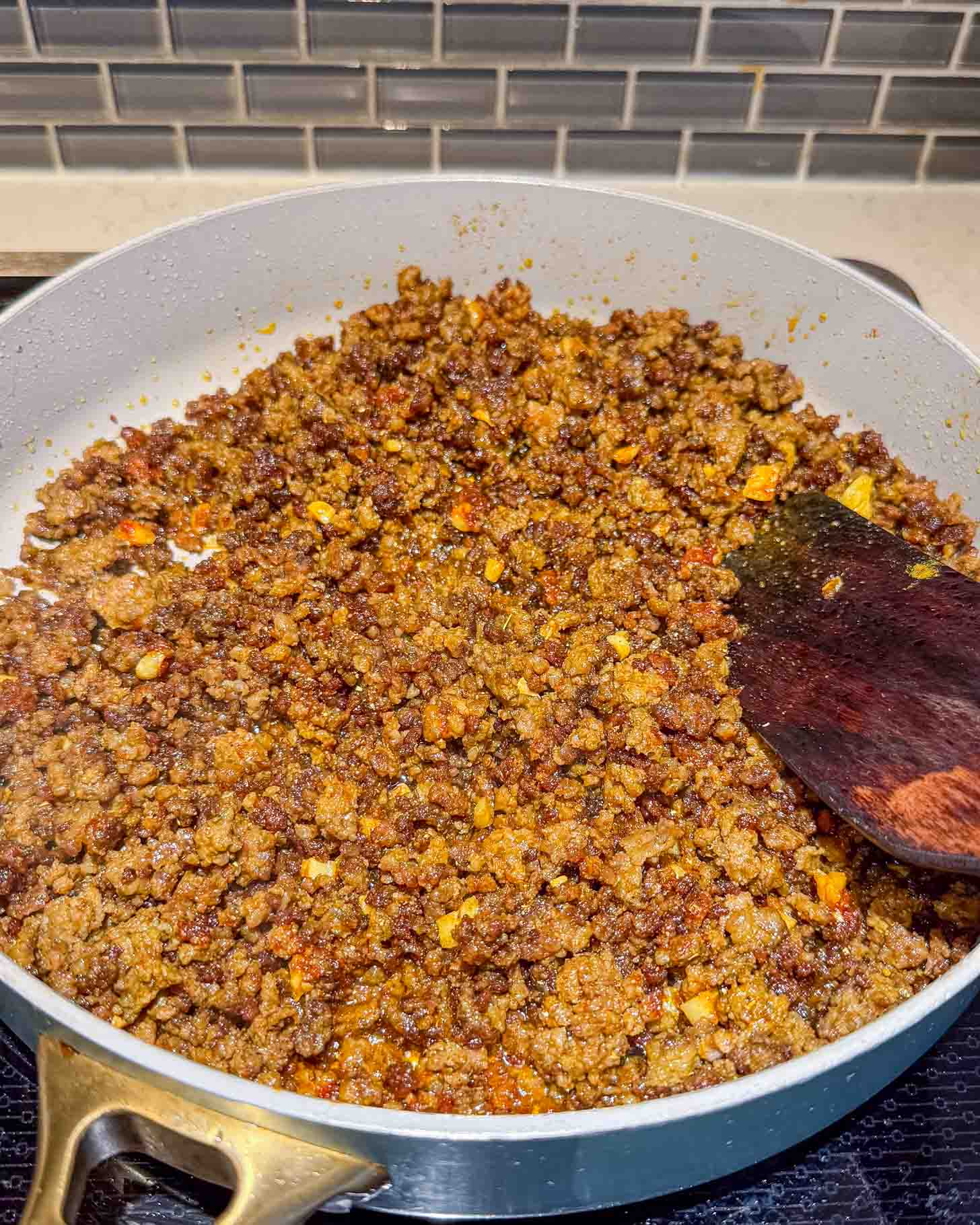 A sauté pan filled with cooked, seasoned ground beef being stirred with a wooden spatula on a stove, with a tile backsplash in the background.
