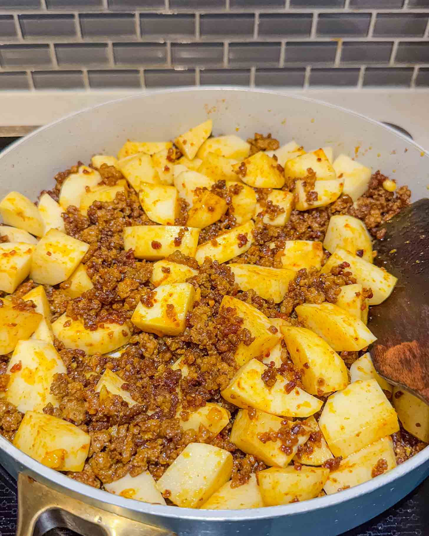 A skillet filled with browned ground beef and cubed potatoes, seasoned and cooking on a stovetop, with a wooden spatula resting on the side. Gray subway tile backsplash is visible in the background.