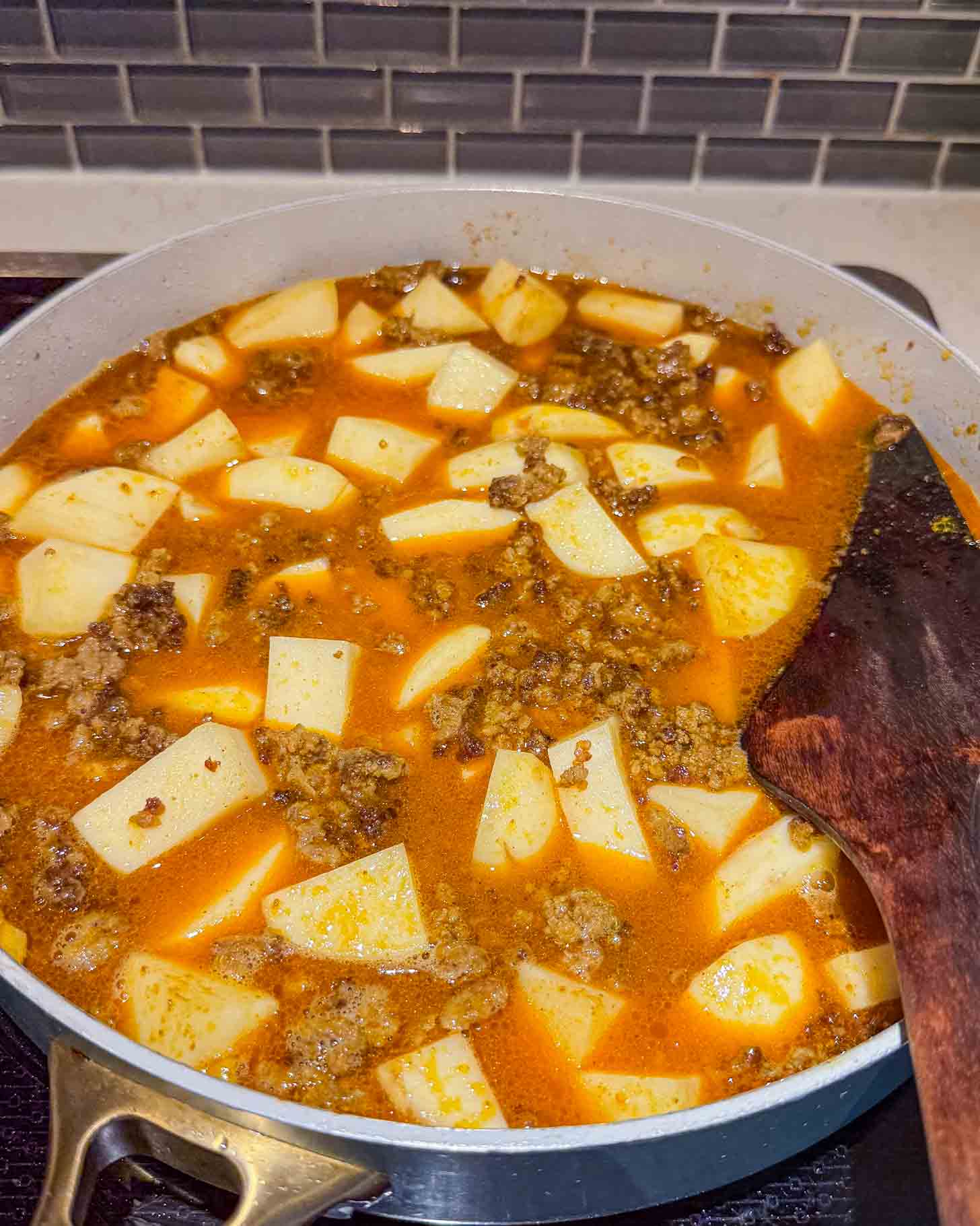 A pot filled with a stew of ground meat, diced potatoes, and a red-orange broth is cooking on a stovetop. A wooden spatula rests on the side of the pot.