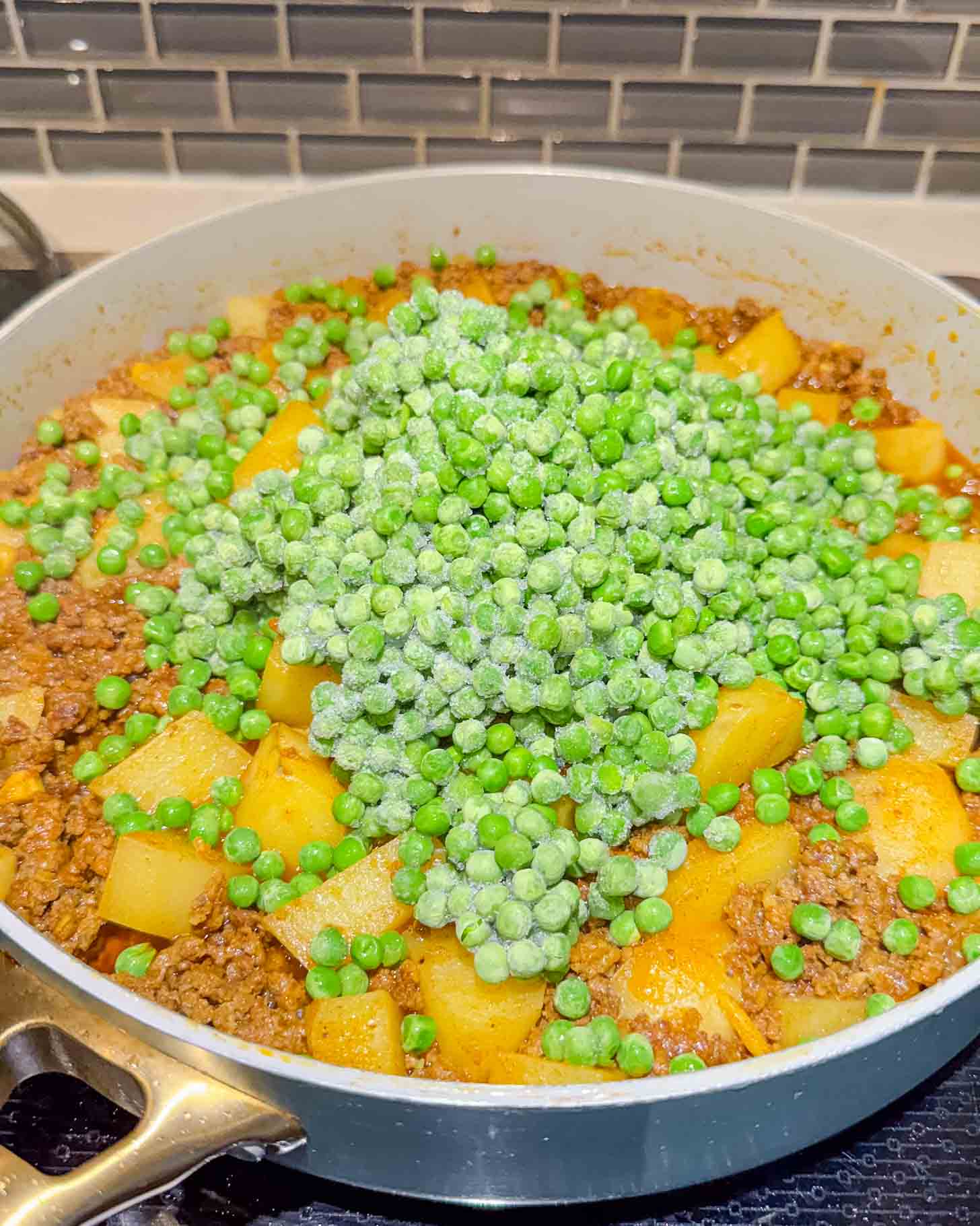 A pan filled with ground meat, diced potatoes, and a pile of frozen green peas, with a gray tile backsplash in the background.