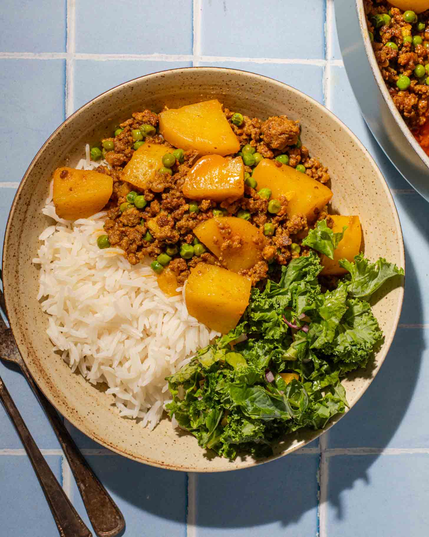 A bowl containing white rice, ground meat with potatoes and peas in sauce, and a side of fresh green salad, on a blue tiled surface.