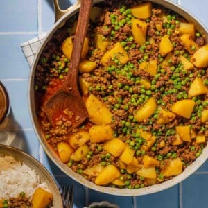 A skillet filled with ground meat, potatoes, peas, and spices sits on a blue tiled surface, with a wooden spoon resting inside. Nearby are a bowl of rice and a small dish of chopped herbs.
