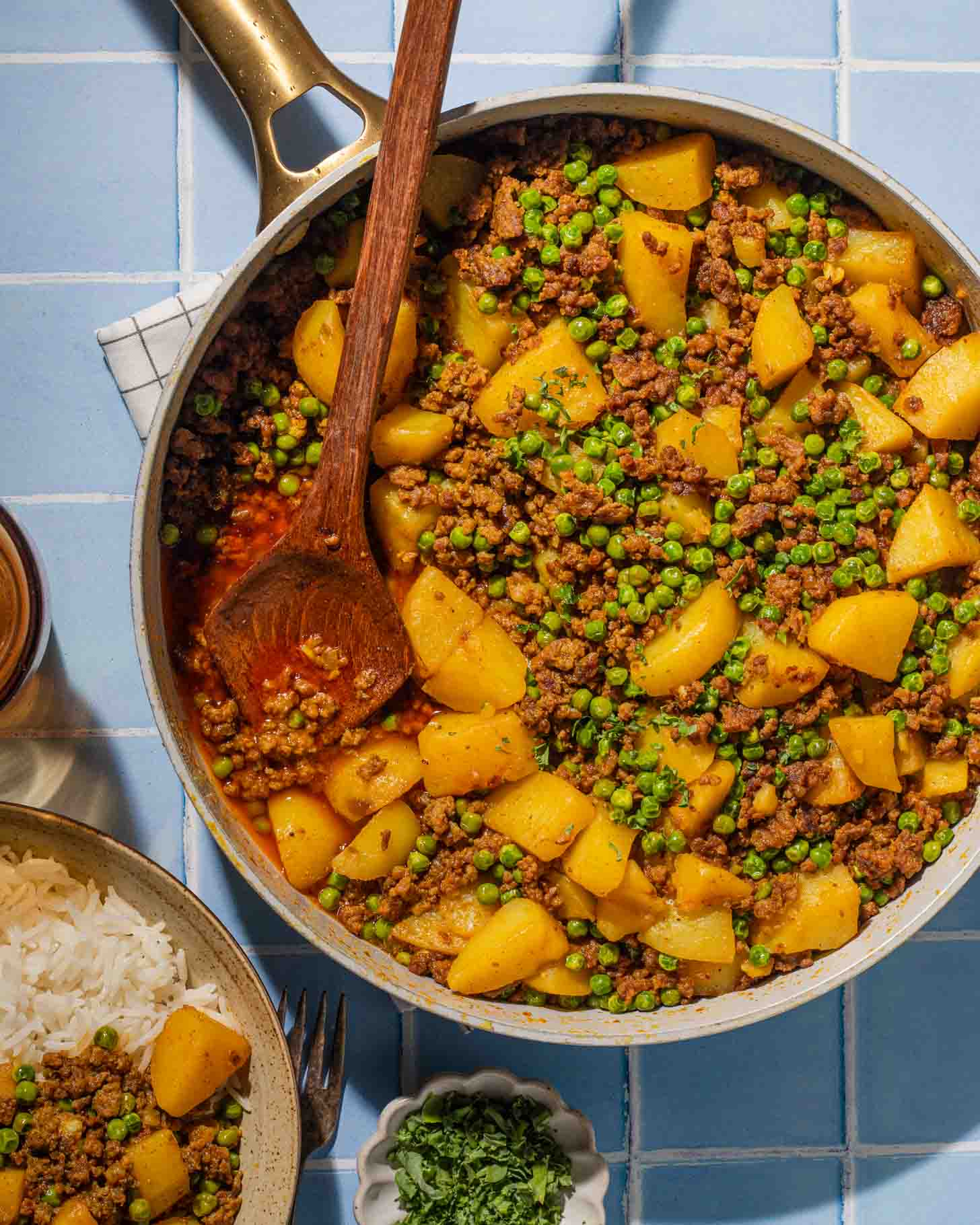 A skillet filled with ground meat, potatoes, peas, and spices sits on a blue tiled surface, with a wooden spoon resting inside. Nearby are a bowl of rice and a small dish of chopped herbs.