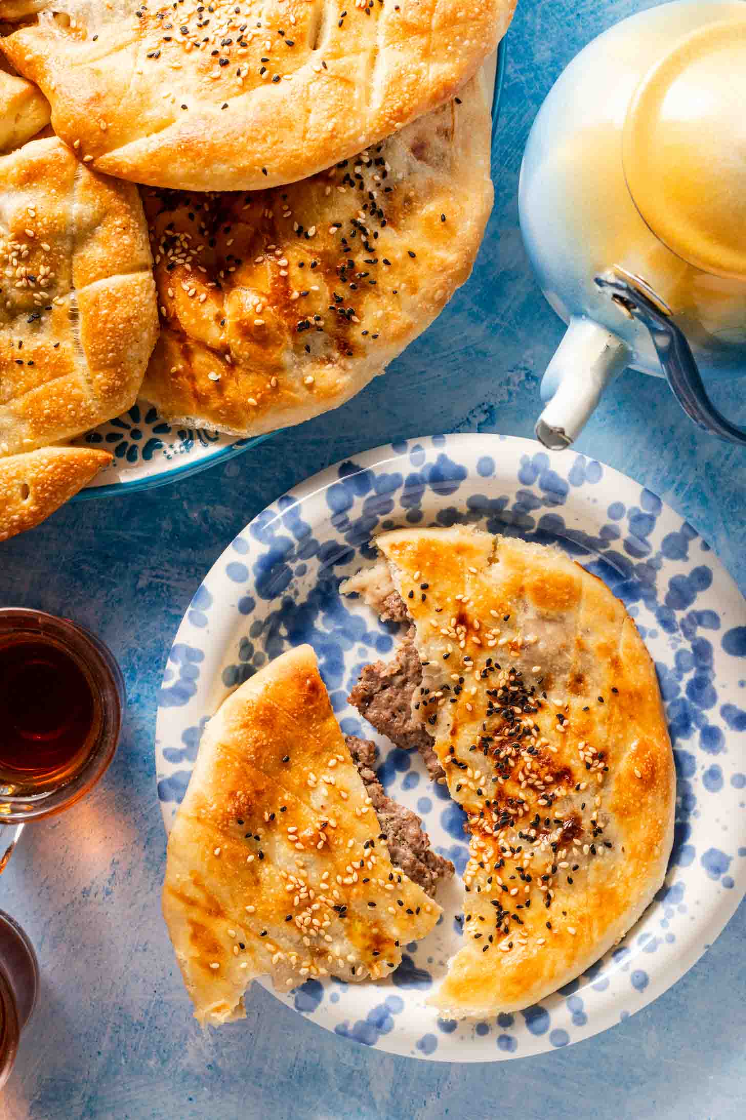 A round flatbread stuffed with meat and topped with sesame seeds sits on a blue-patterned plate. A teapot and cups of tea are nearby, and more bread is on a larger plate in the background.