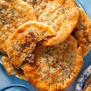 A plate of round flatbreads, some topped with sesame and nigella seeds, one with a bite taken showing a ground meat filling, next to a glass teapot and a tea cup on a blue surface.