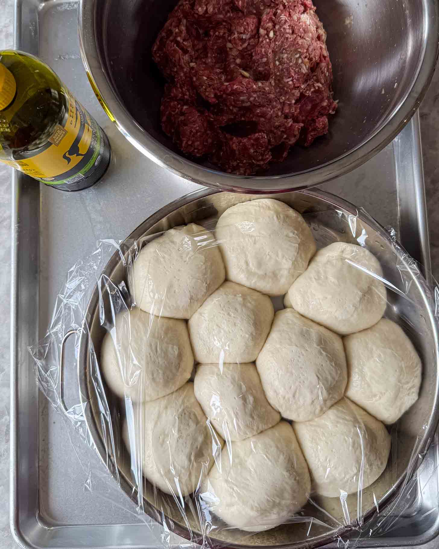 A bowl of raw dough balls covered in plastic wrap, a bowl of seasoned ground meat, and a bottle of olive oil on a metal tray.