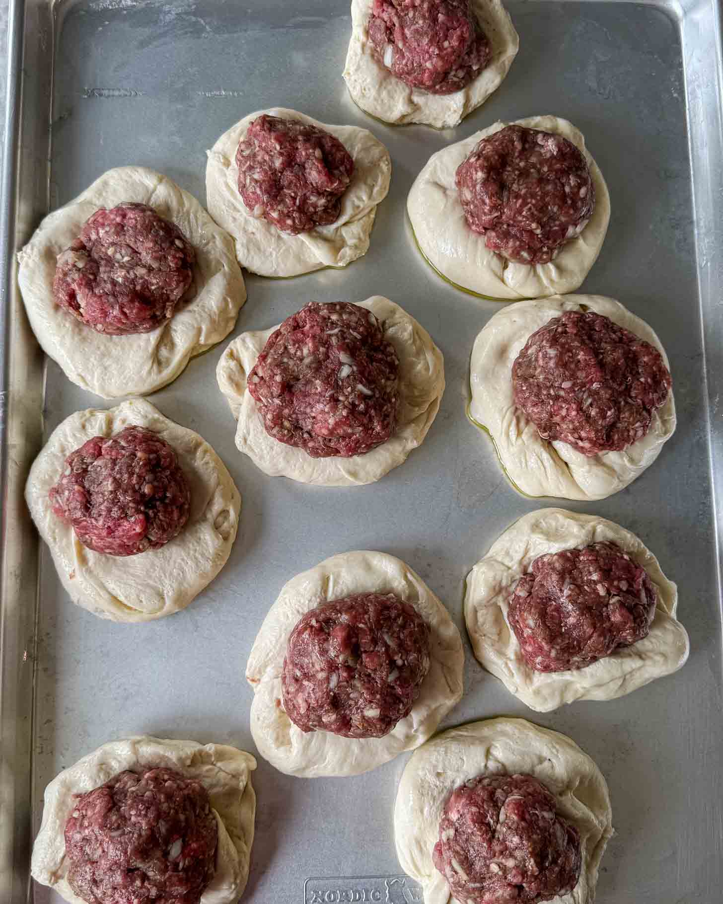 Unbaked dough rounds topped with raw ground meat are arranged on a metal baking sheet, ready to be cooked.