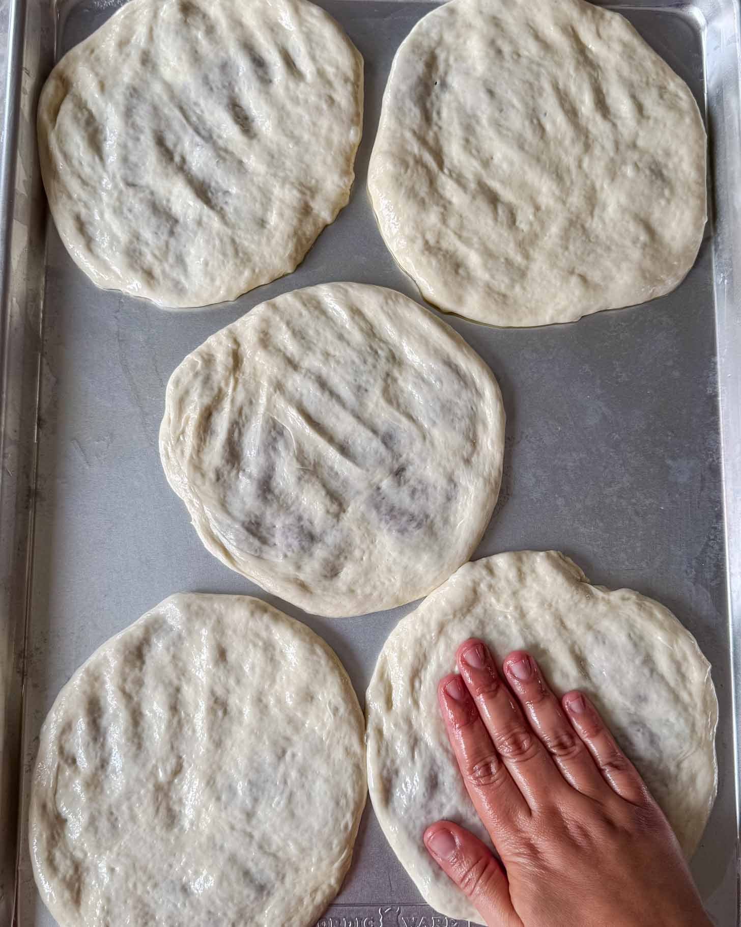 A hand presses and shapes round pieces of raw pizza dough on a metal baking sheet, with five dough circles spread out on the tray.