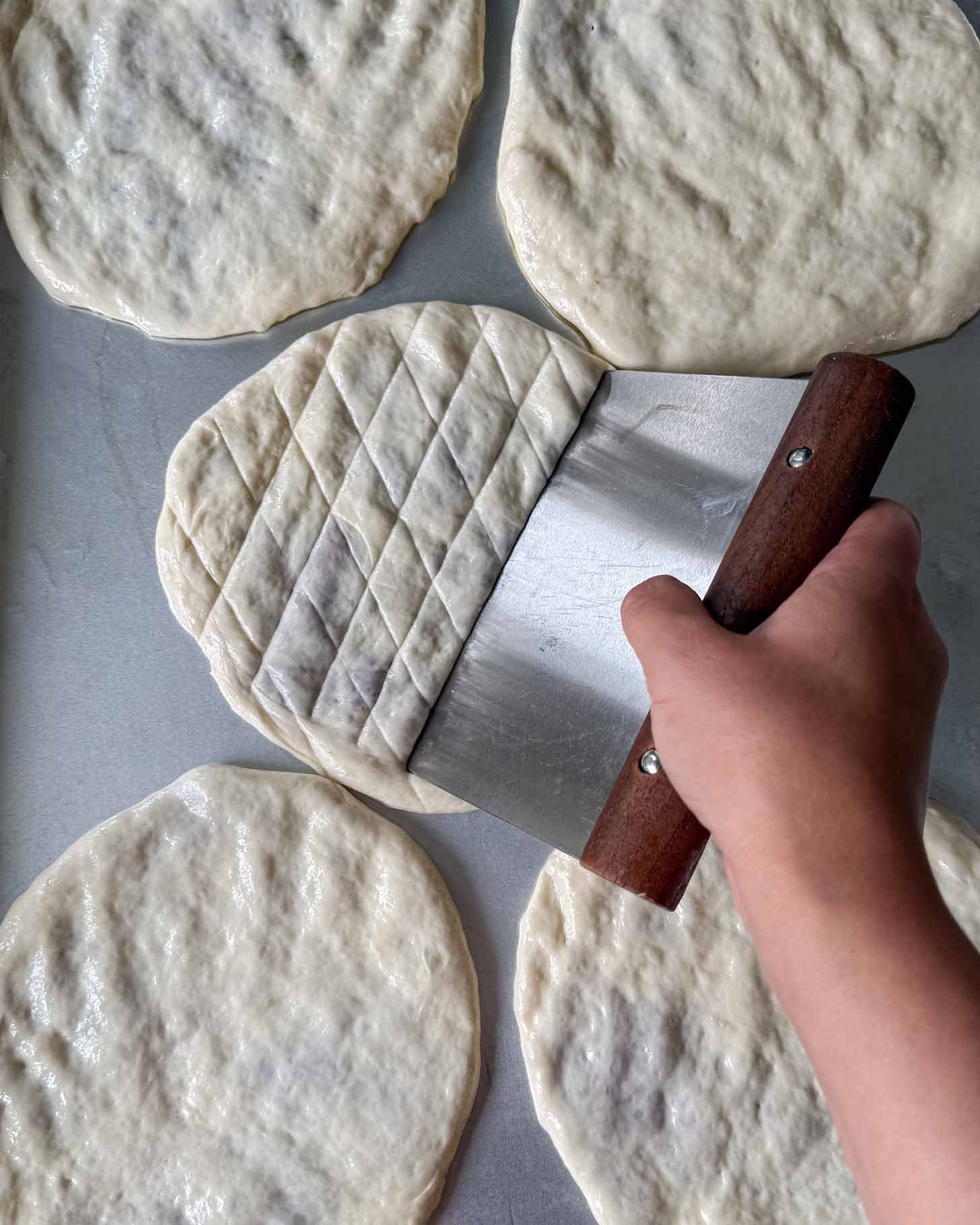 A hand uses a dough scraper to score a diamond pattern onto a piece of round dough among several unscored dough rounds on a metal surface.