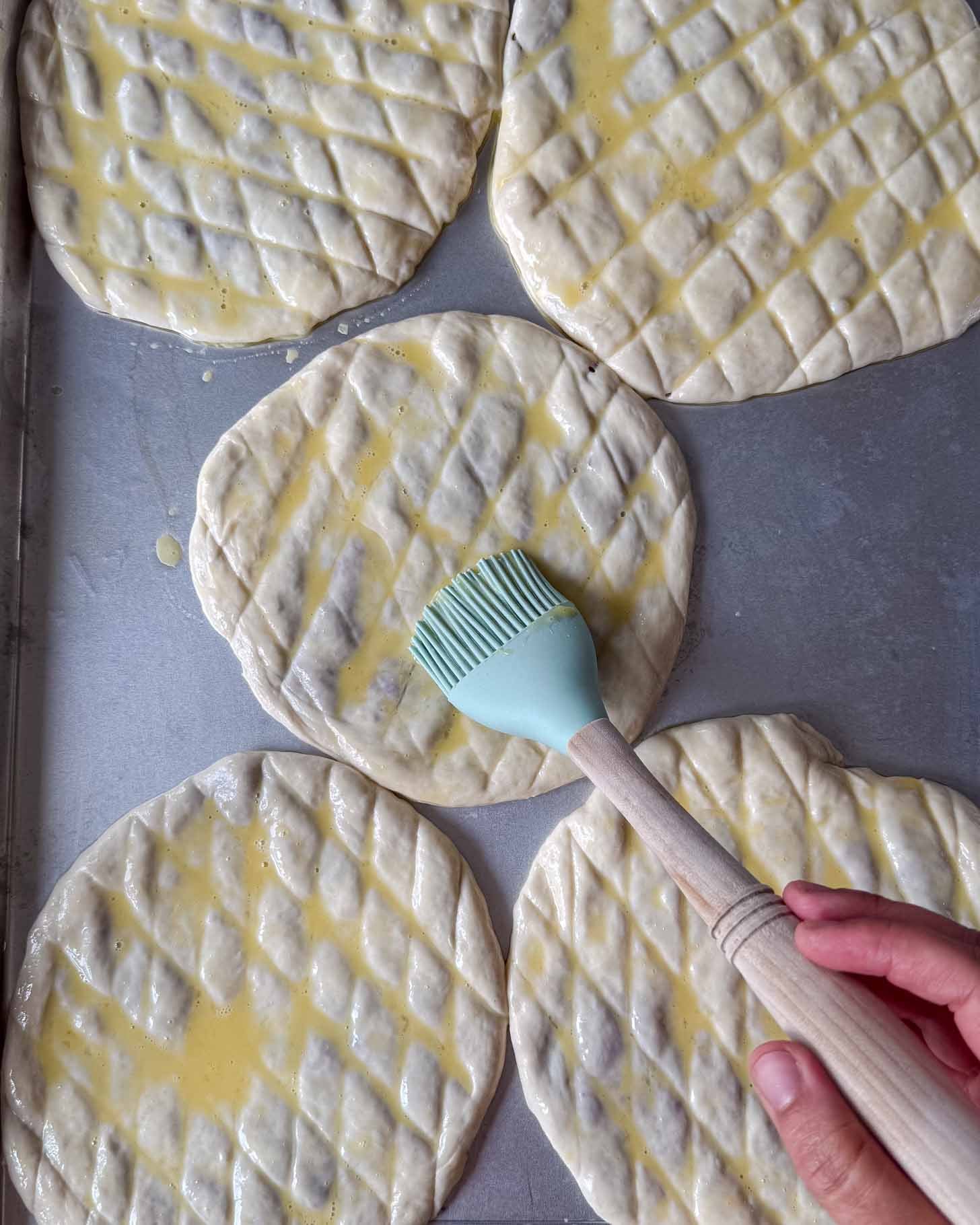 Five round pieces of dough with a diamond pattern are brushed with egg wash by a hand holding a silicone pastry brush on a metal baking sheet.