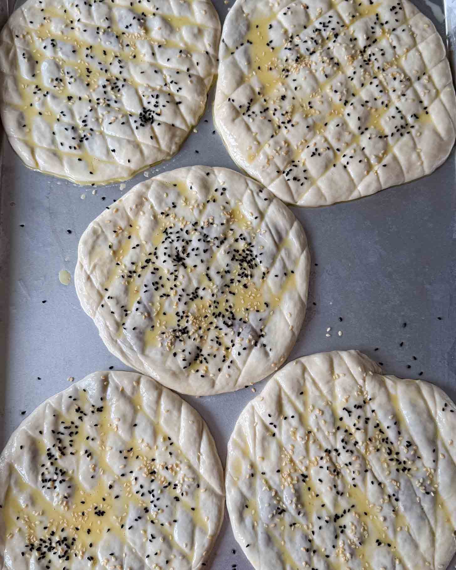 Five round, uncooked flatbreads with a crosshatch pattern, brushed with egg wash and sprinkled with black and white sesame seeds, are arranged on a metal baking tray.
