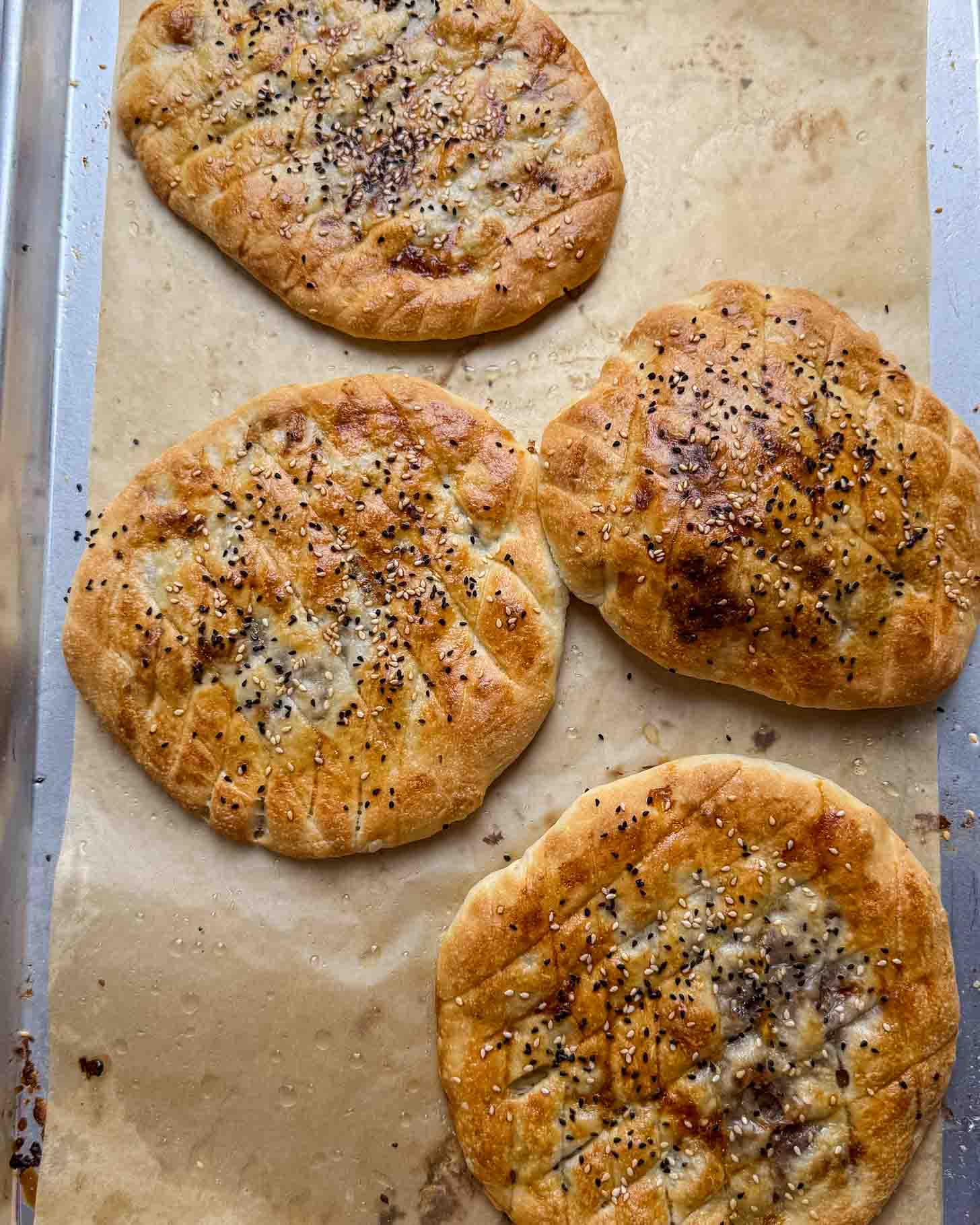 Four round, golden brown breads with sesame and black seeds on top, arranged on parchment paper on a baking tray. The breads have a crisscross pattern on the surface.