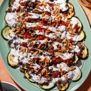 A platter of roasted zucchini slices topped with ground meat, tomato sauce, pine nuts, and a drizzle of white yogurt sauce, set on a peach-colored tiled surface.