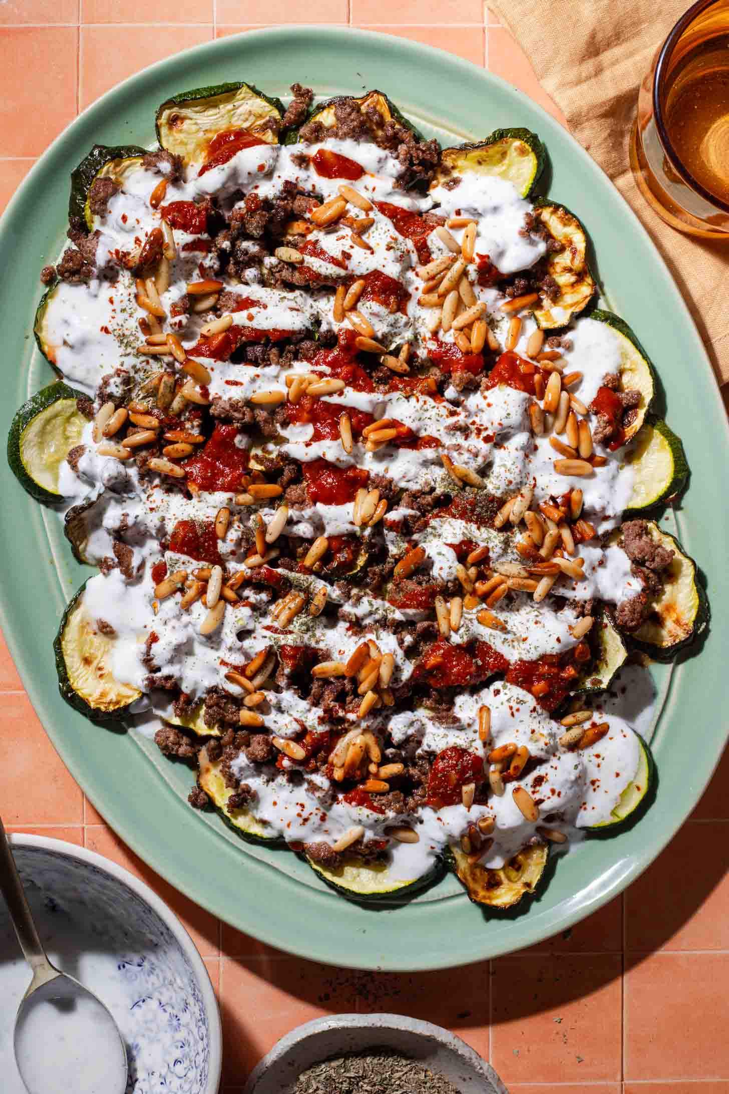 A platter of roasted zucchini slices topped with ground meat, tomato sauce, pine nuts, and a drizzle of white yogurt sauce, set on a peach-colored tiled surface.