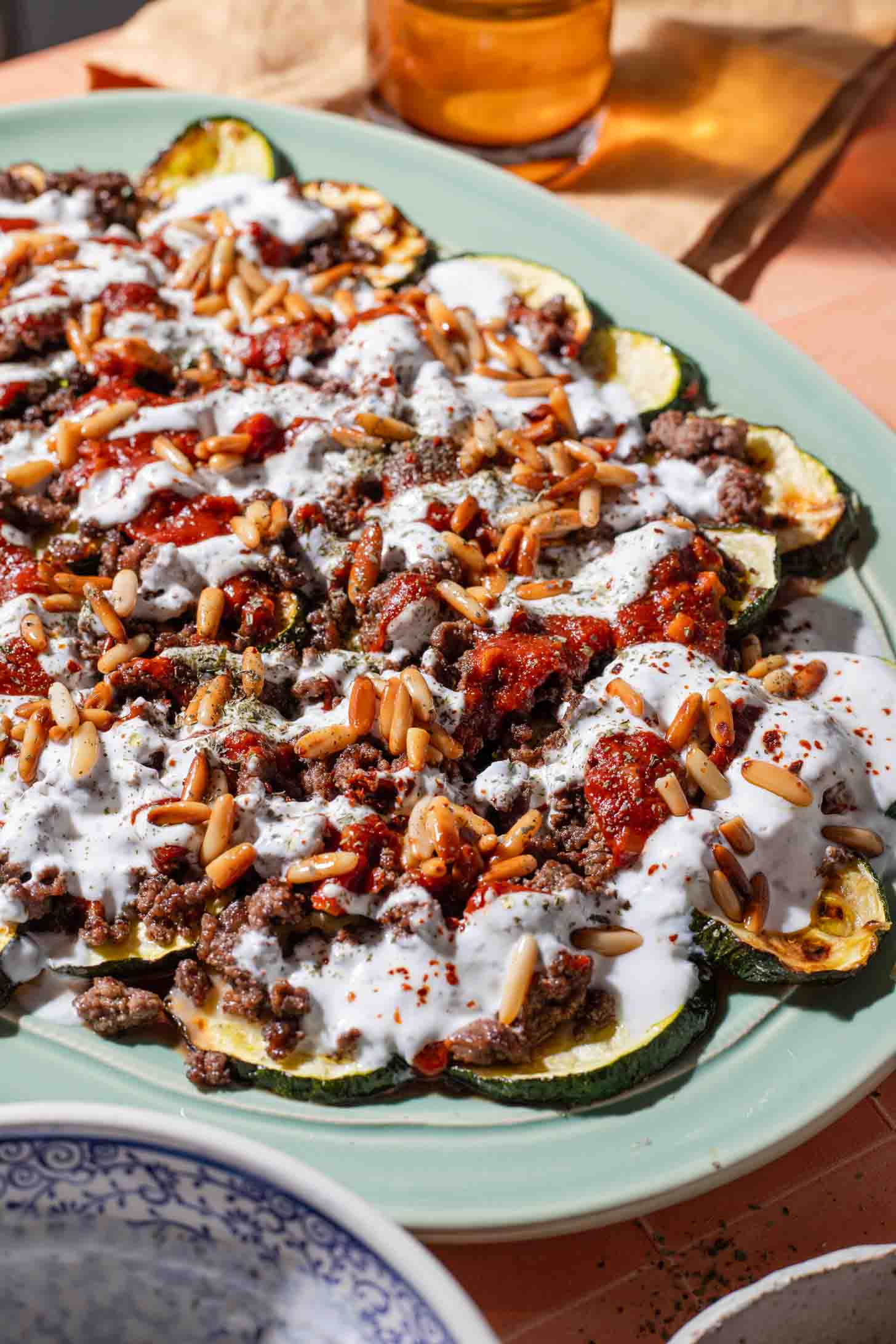 A plate of roasted zucchini rounds topped with ground meat, tomato sauce, yogurt sauce, toasted pine nuts, and herbs on a light green dish.