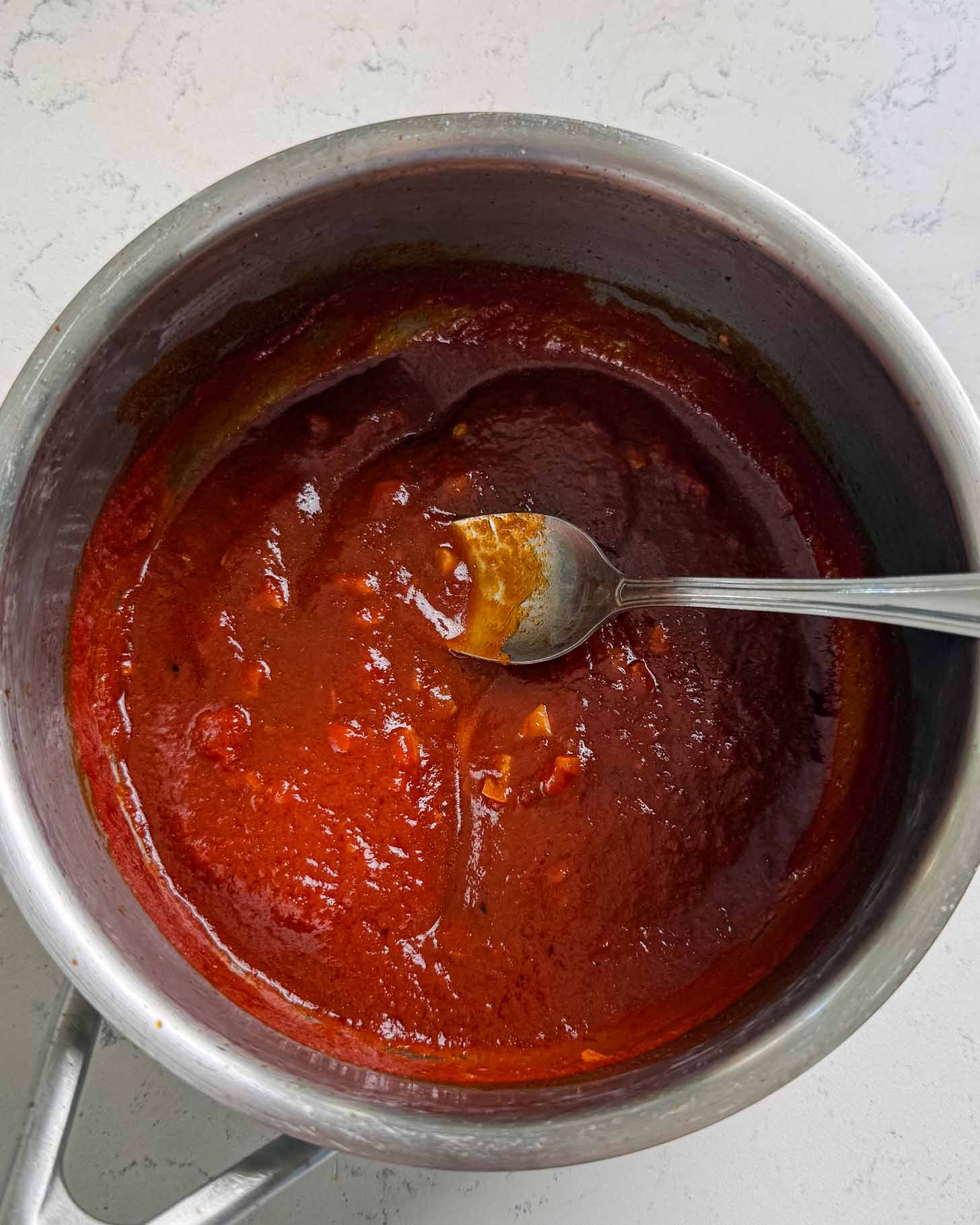A pot of thick red barbecue sauce with a silver spoon resting inside, placed on a white countertop.