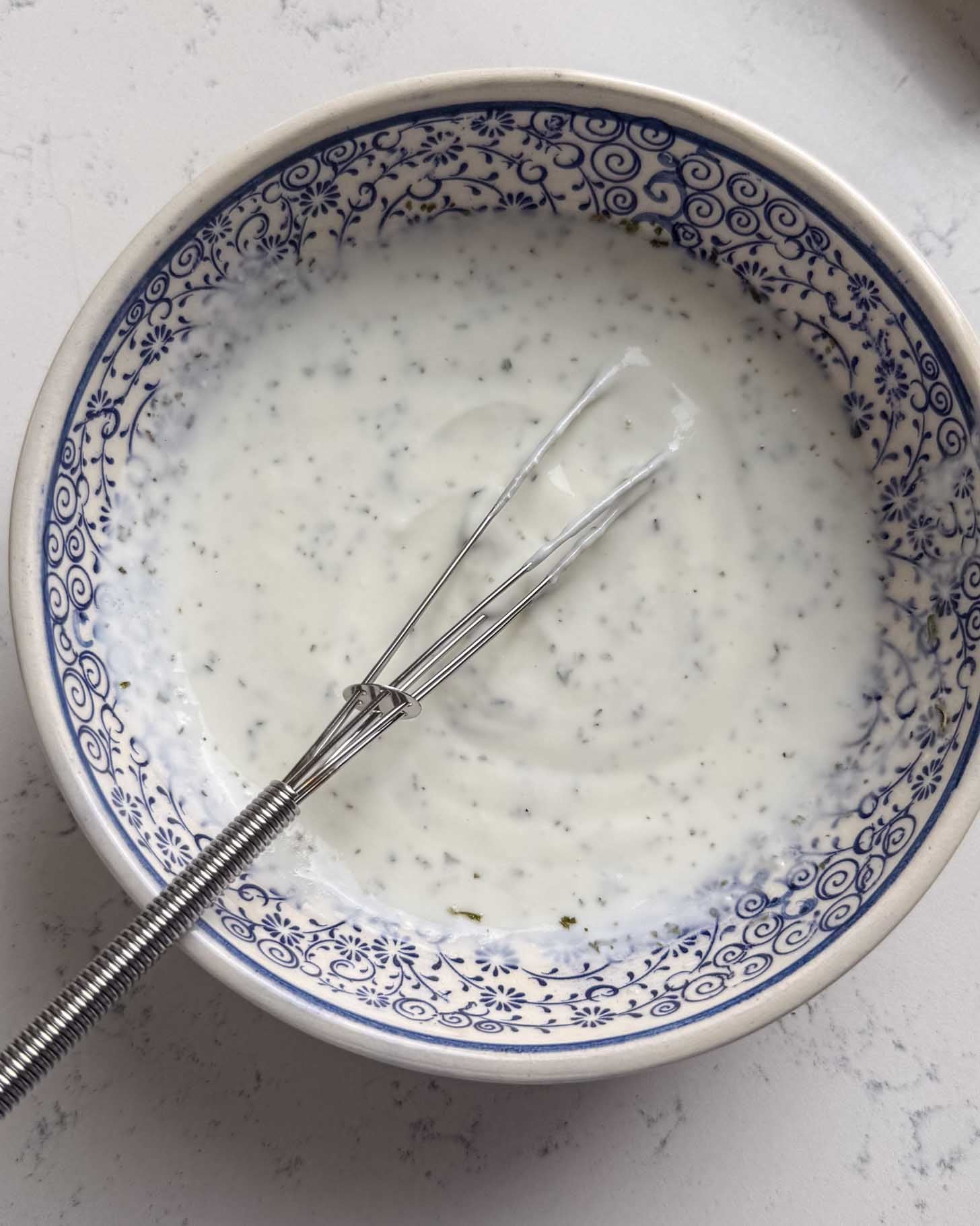 A bowl with blue patterns filled with creamy white sauce or dressing, with visible herbs, and a small metal whisk resting inside the bowl.