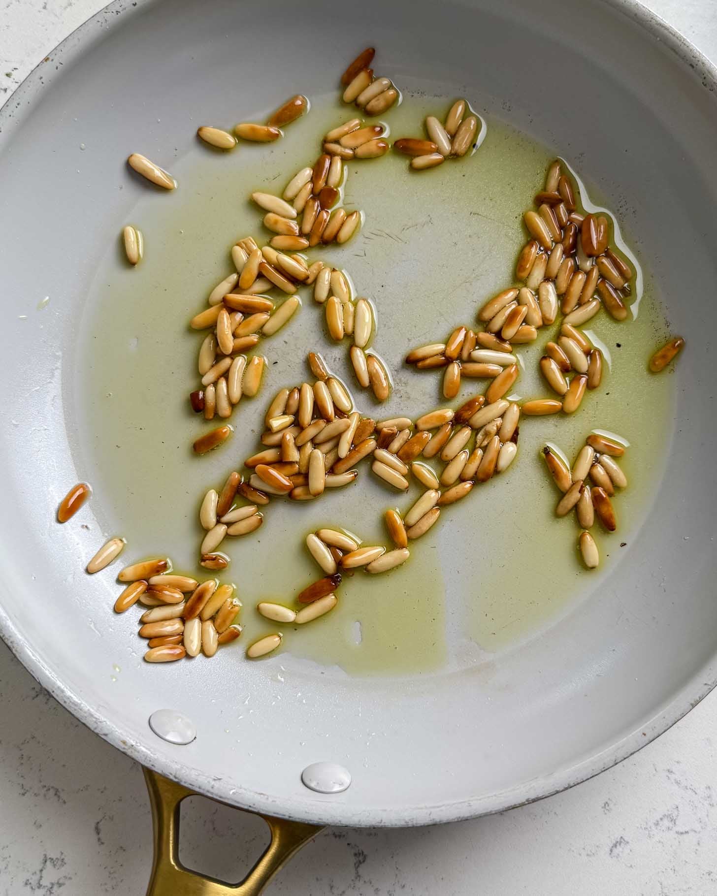 Pine nuts toasting in olive oil in a white skillet with a gold handle, some nuts turning golden brown.