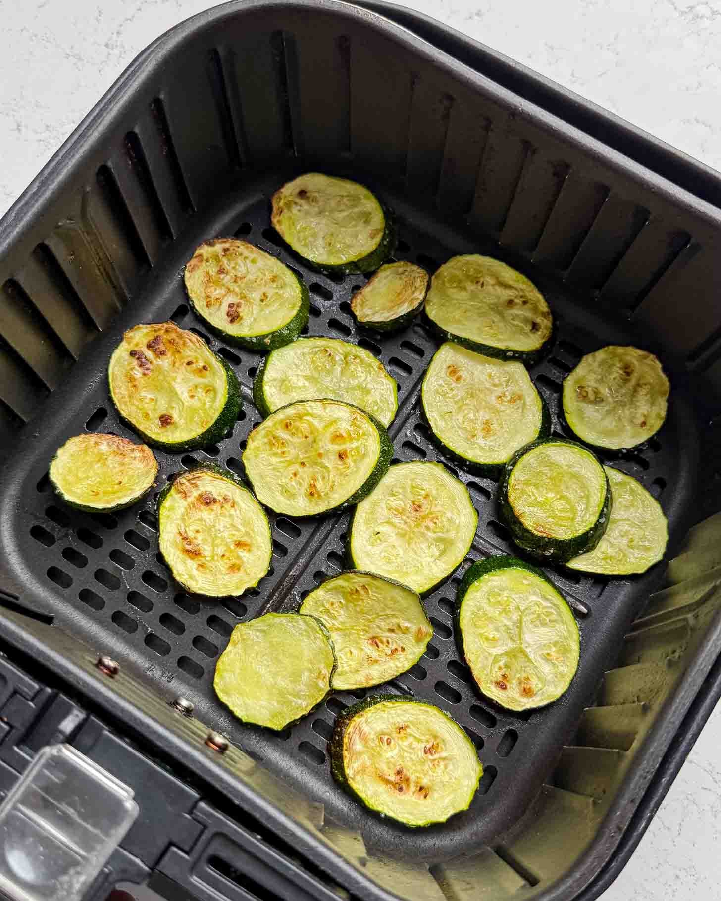 Sliced zucchini rounds, lightly browned, are arranged in a single layer inside an air fryer basket on a white countertop.