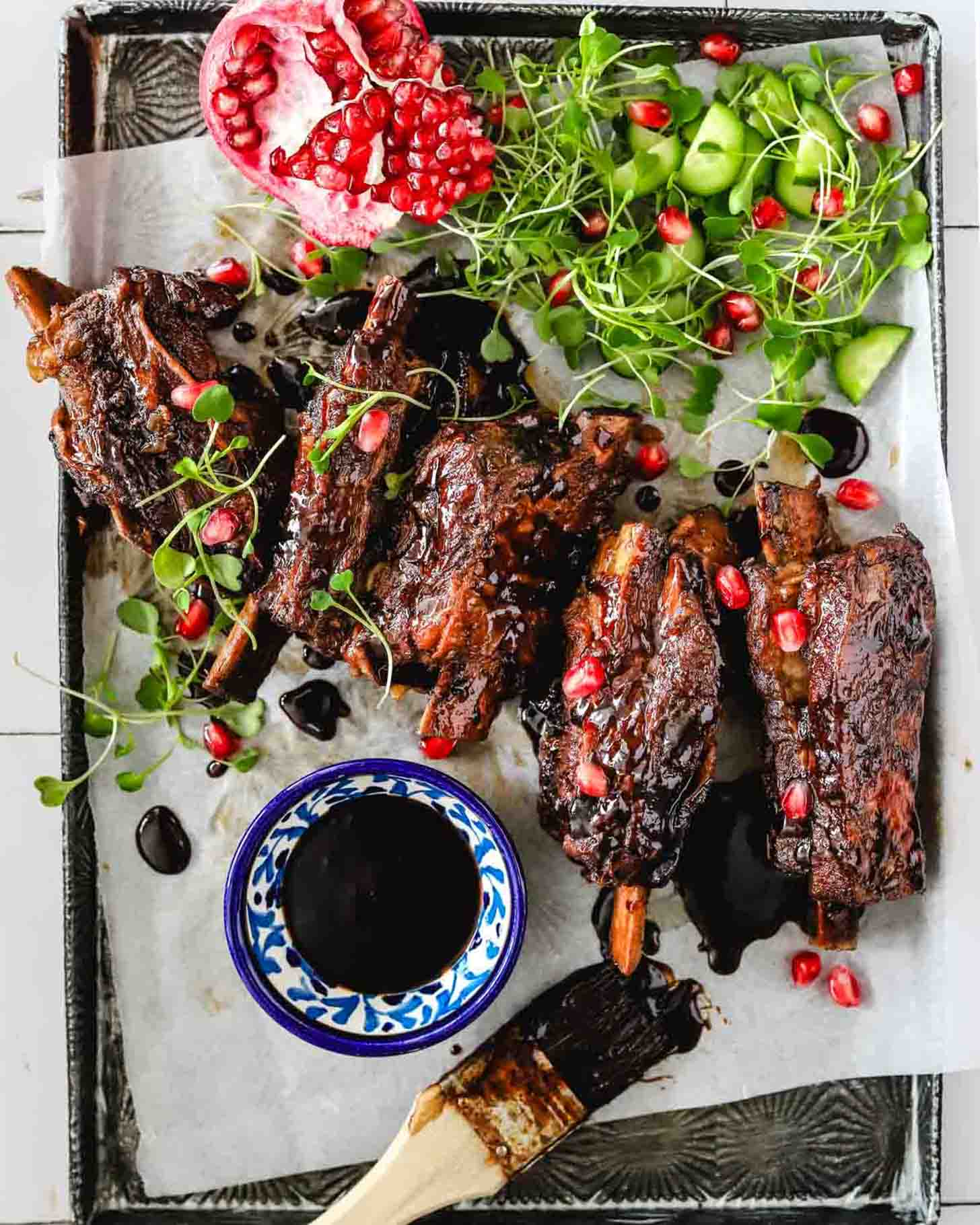 Barbecued ribs garnished with microgreens and pomegranate seeds, served with a small bowl of sauce, a brush, cucumber pieces, and a halved pomegranate on a tray lined with parchment paper.