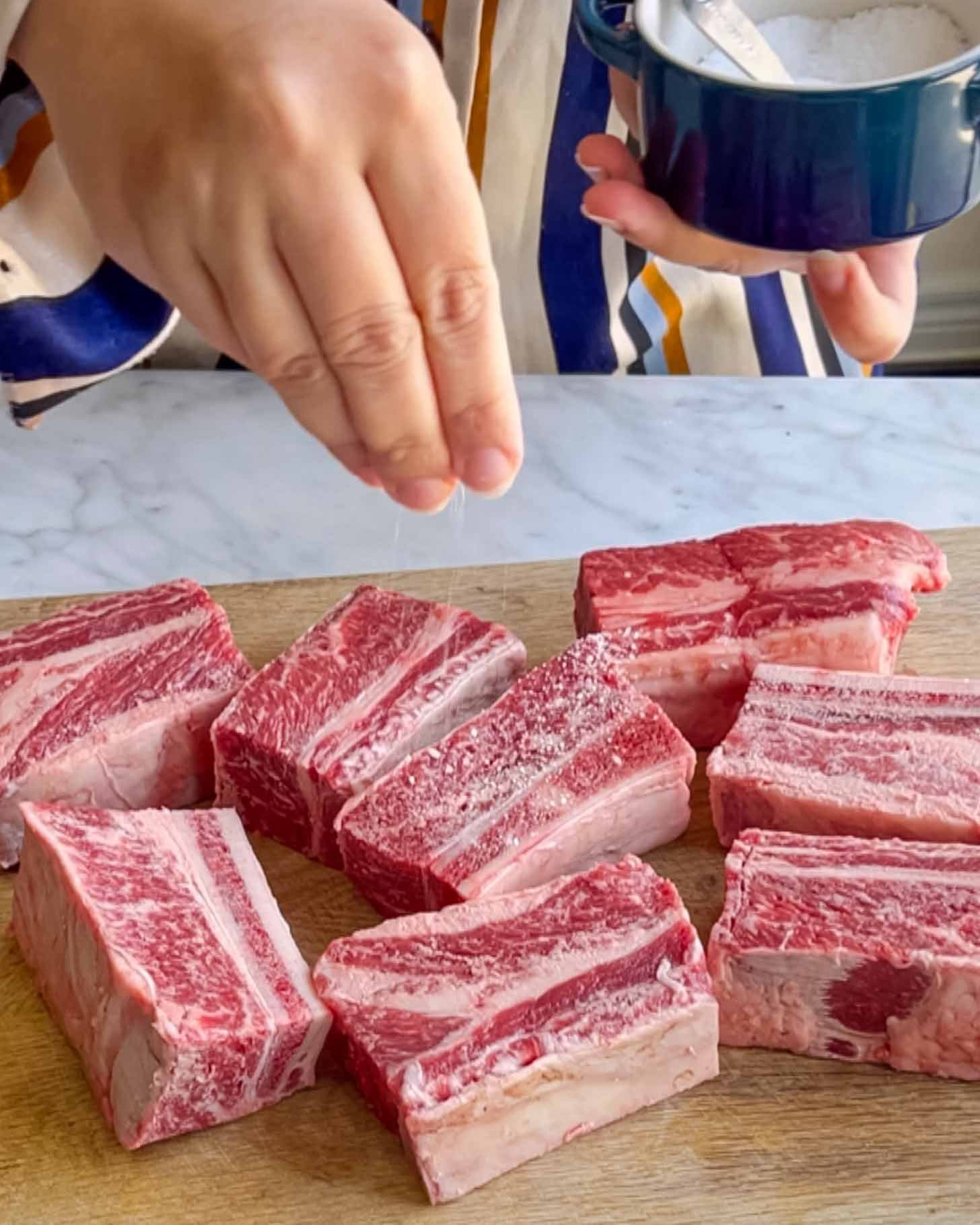 A person sprinkles salt onto several pieces of raw beef short ribs arranged on a wooden cutting board.