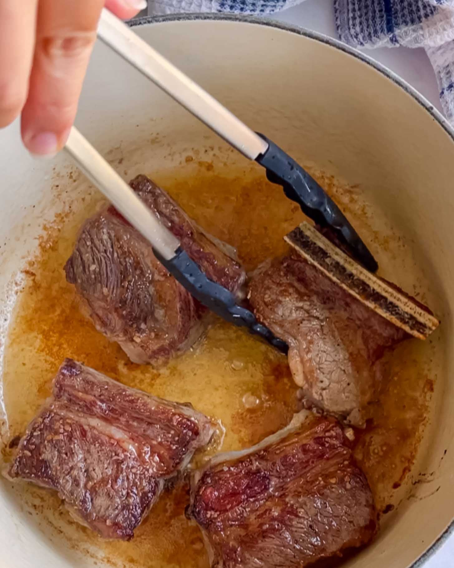 A hand uses tongs to sear short ribs in a pot with oil. The meat is browning on all sides, creating a crisp exterior.