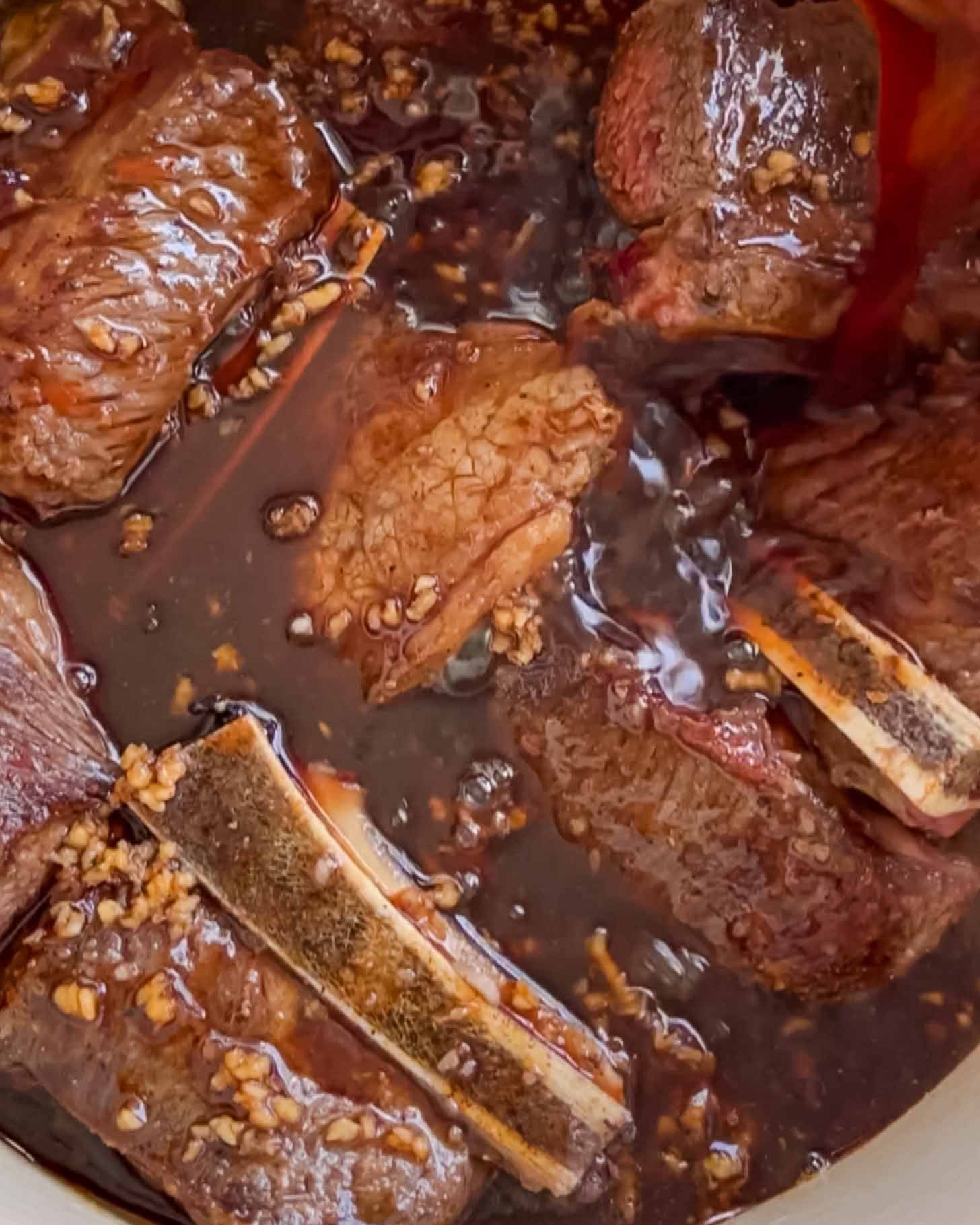 Chunks of beef short ribs with bones simmer in a dark, rich sauce with visible minced garlic, creating a hearty, savory dish.