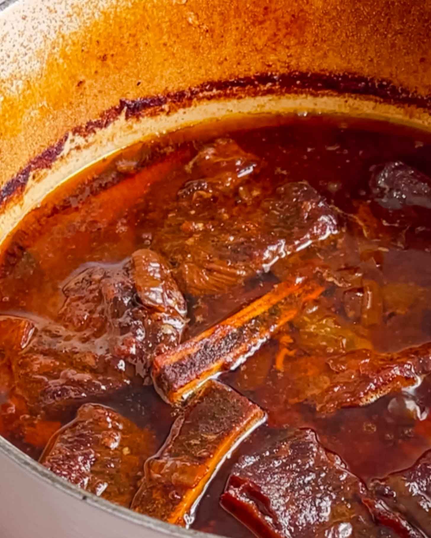 Chunks of beef short ribs simmer in a rich, dark brown sauce inside a large pot with browned edges, indicating slow cooking and tender meat.