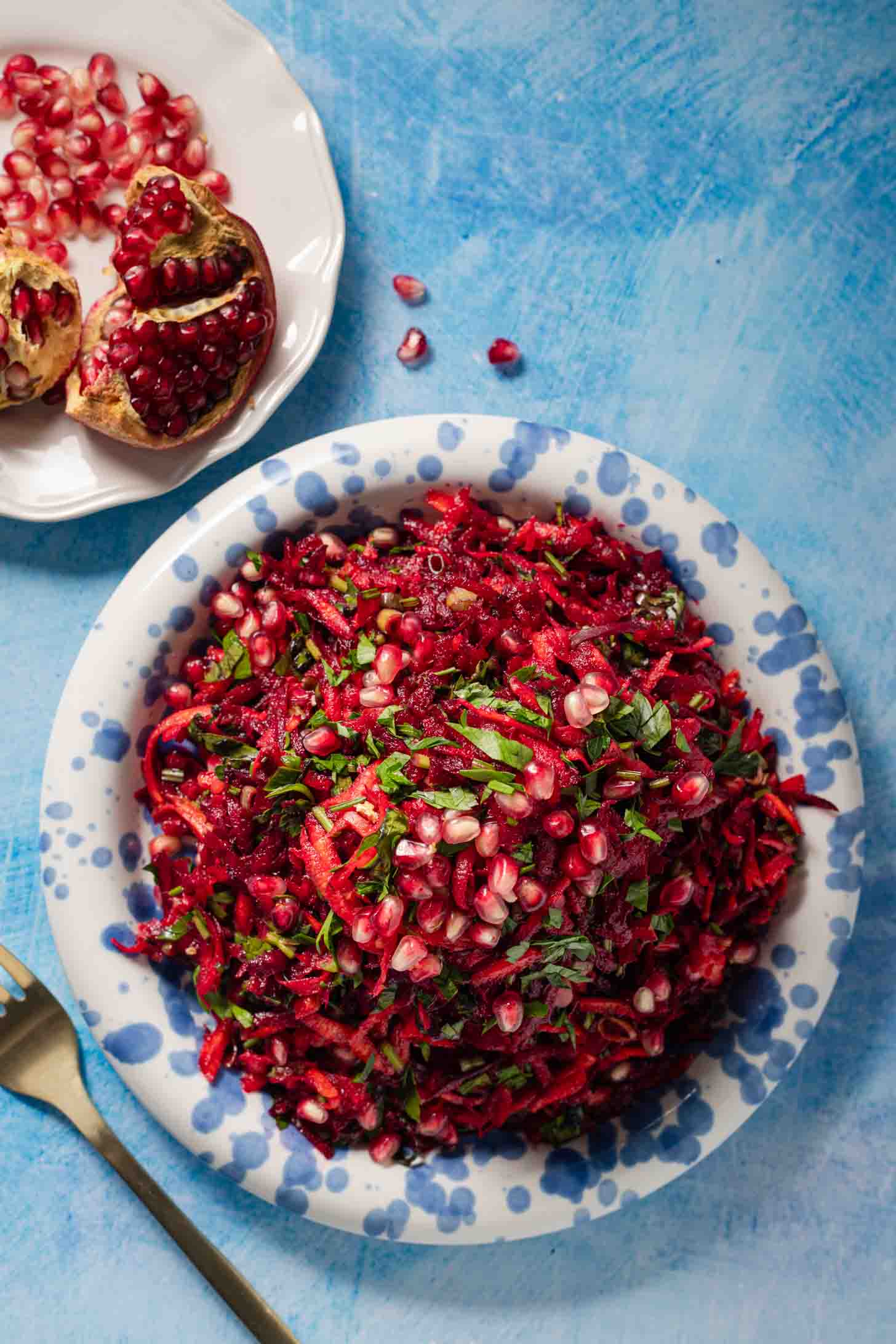 A plate of shredded beet and carrot salad topped with pomegranate seeds and herbs sits on a blue background. A fork and a halved pomegranate are beside the plate.