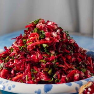 A plate piled with shredded beet salad, garnished with fresh herbs and pomegranate seeds, sits on a blue surface. A pomegranate half is visible in the foreground.